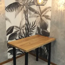 Drewniany blat stołu z czarnymi nogami, oświetlony wiszącą lampą w stylu loft, na tle ściany z motywem tropikalnych liści i marmurowej podłogi.