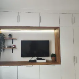 Białe szafki z drewnianą wnęką na telewizor, w której znajdują się półki z dekoracjami i oświetlenie LED.