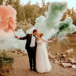 Para młoda całuje się, trzymając w dłoniach race dymne w kolorach koralowym i zielonym, na tle jeziora i drzew podczas sesji ślubnej.