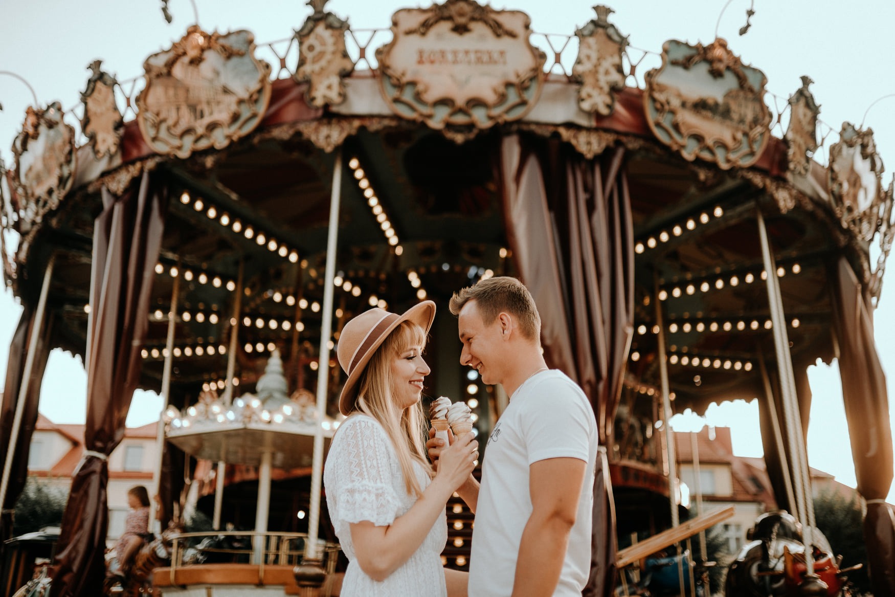 Uśmiechnięta para trzymająca lody na tle karuzeli w stylu vintage z dekoracyjnymi zdobieniami i ciepłym oświetleniem.