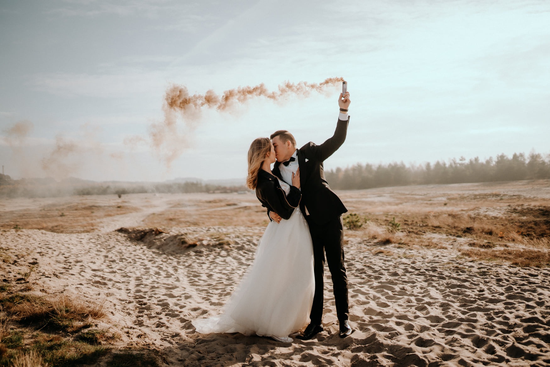 Para młoda całuje się na piaszczystej wydmie, pan młody trzyma w górze odpaloną świecę dymną brązowego koloru, w tle widok na zalesiony teren pod błękitnym niebem.
