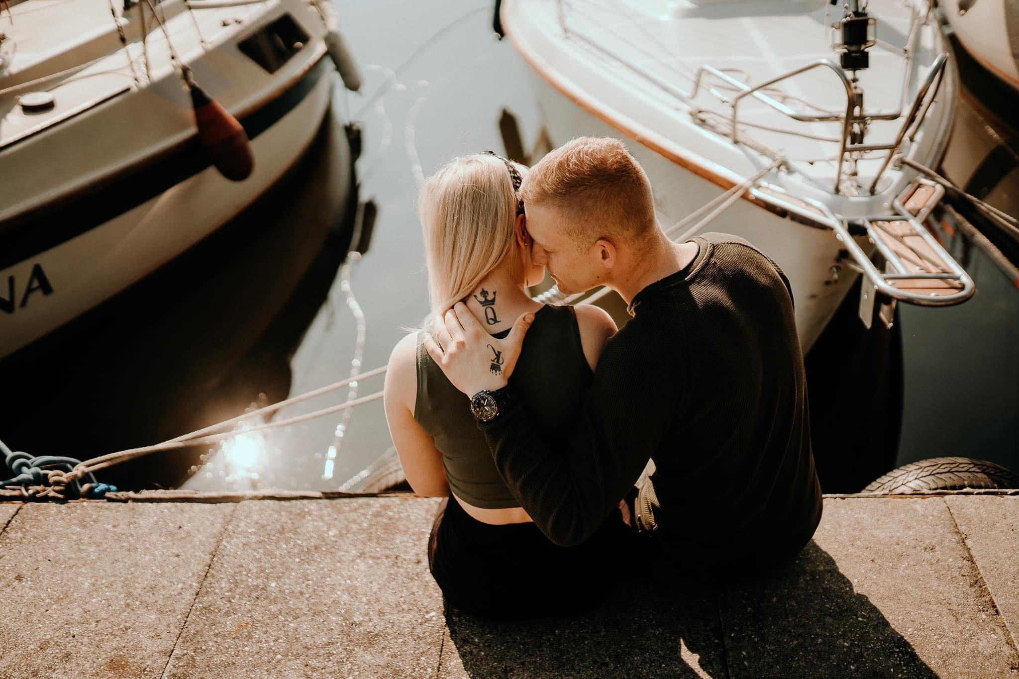 Para zakochanych siedzi na nabrzeżu portowym, przytulona do siebie na tle dwóch jachtów; ona ma tatuaże króla i królowej na karku i dłoni, a on zegarek na nadgarstku.