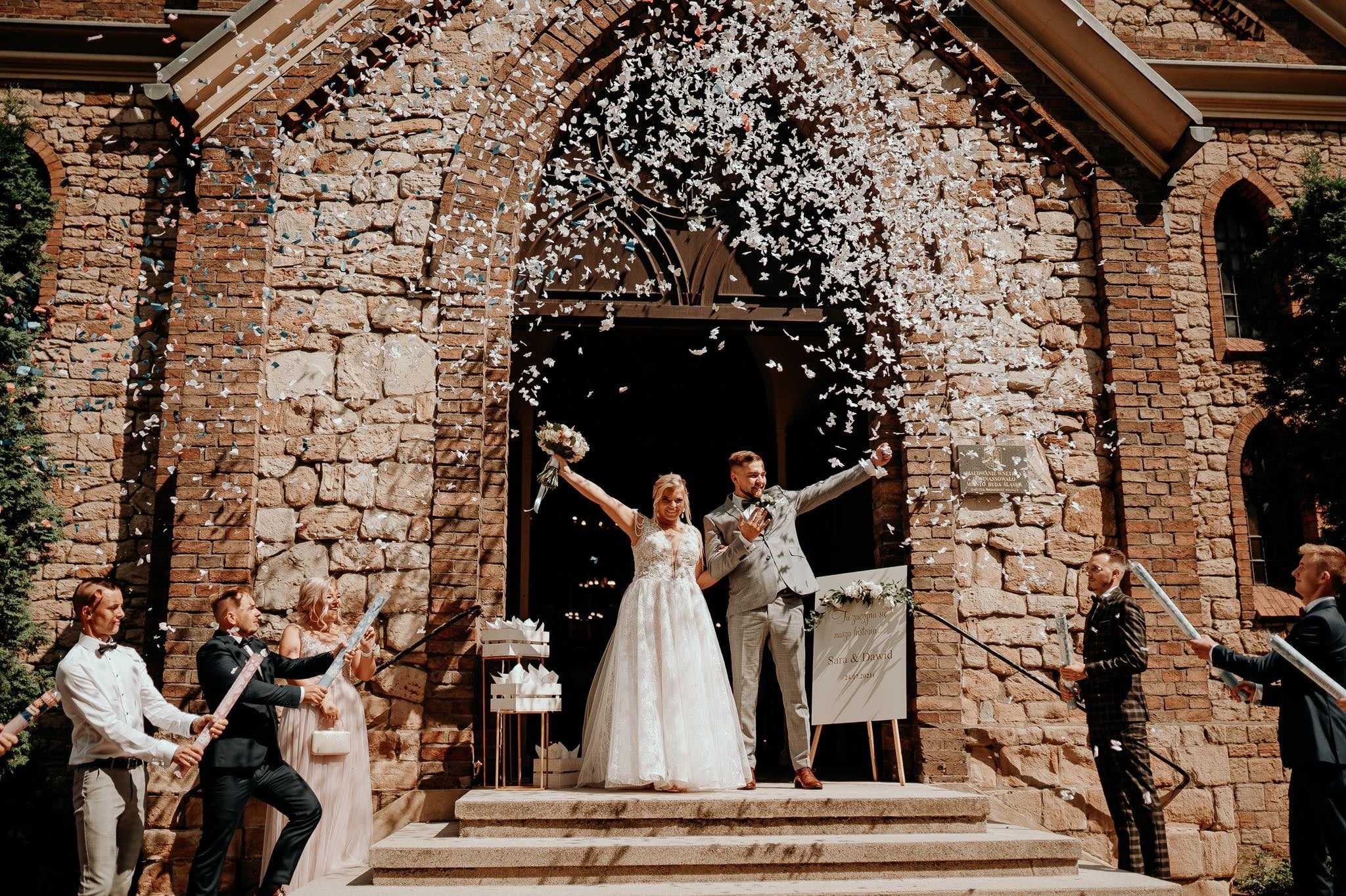 Nowożeńcy opuszczają kościół w Chorzowie, obsypywani konfetti przez gości, widoczni również asystenci z tubami konfetti.