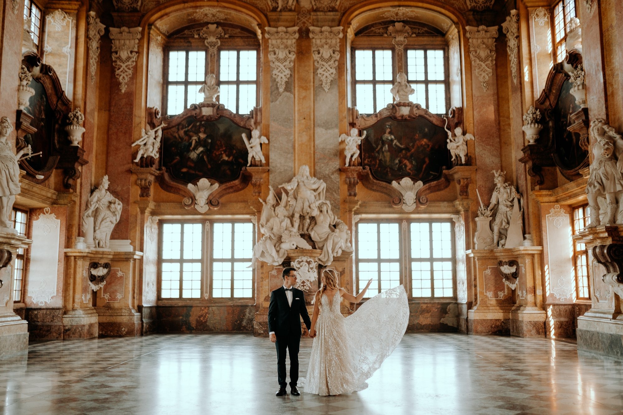 Para młoda trzymająca się za ręce w bogato zdobionym barokowym wnętrzu z rzeźbami i obrazami, panna młoda w sukni z trenem.