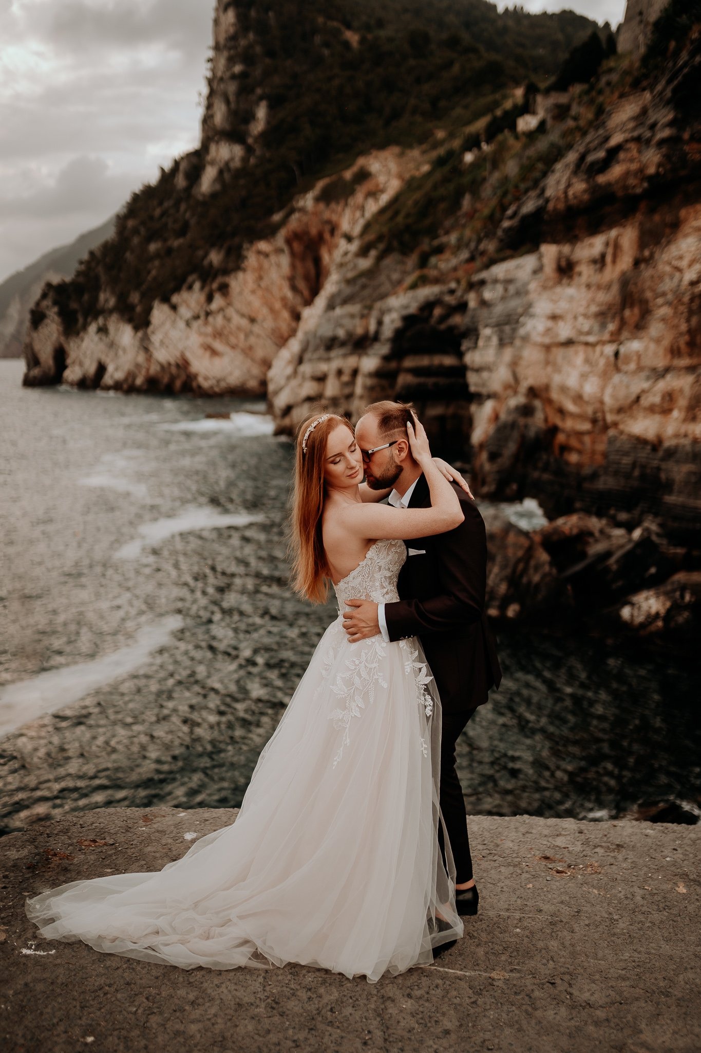 Para młoda w objęciach na tle skalistego wybrzeża i morza, panna młoda w sukni z koronkowymi aplikacjami, pan młody w ciemnym garniturze całuje pannę młodą w skroń.
