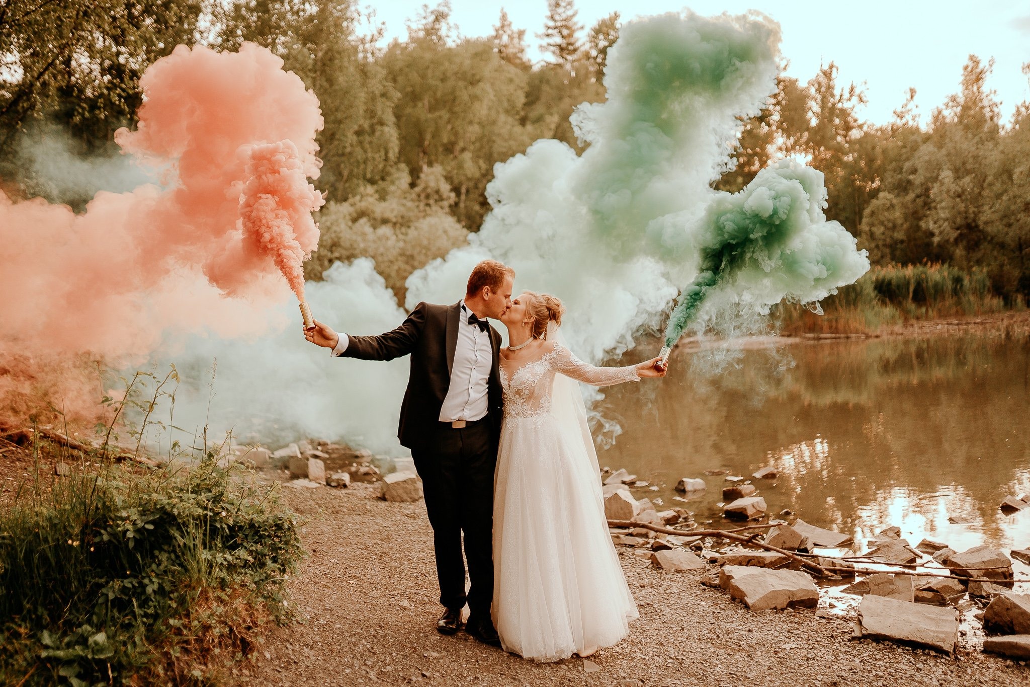 Para młoda całuje się, trzymając w dłoniach race dymne w kolorach koralowym i zielonym, na tle jeziora i drzew podczas sesji ślubnej.
