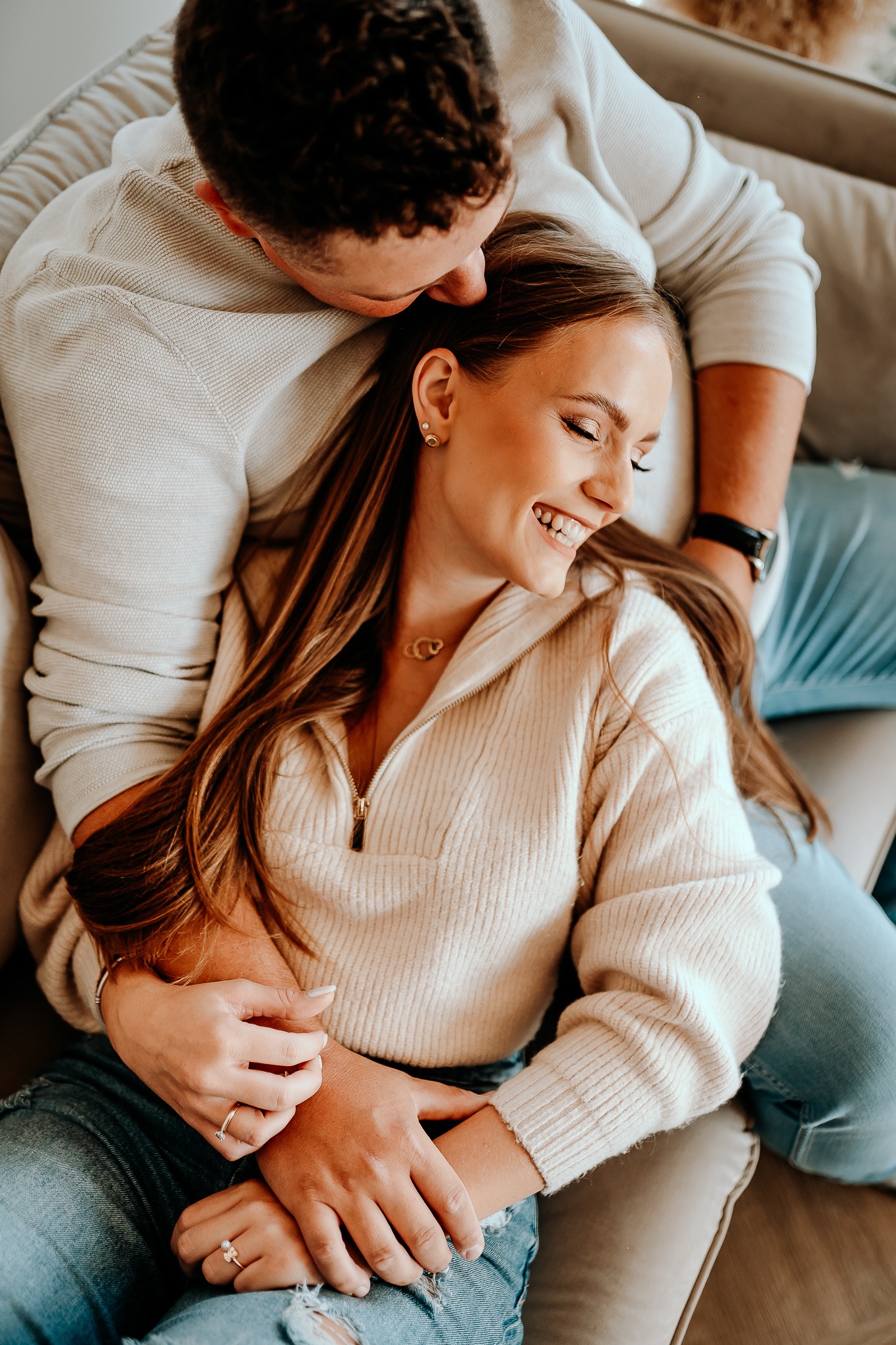 Uśmiechnięta kobieta w beżowym swetrze, obejmowana przez mężczyznę w jasnym swetrze i dżinsach, para siedzi na beżowej sofie, widoczne dłonie z pierścionkami i złoty łańcuszek