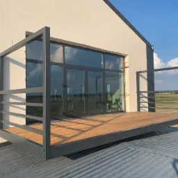 Nowoczesny balkon z drewnianą podłogą i szarą metalową konstrukcją, przylegający do białego budynku z dużymi oknami, widok z dachu.