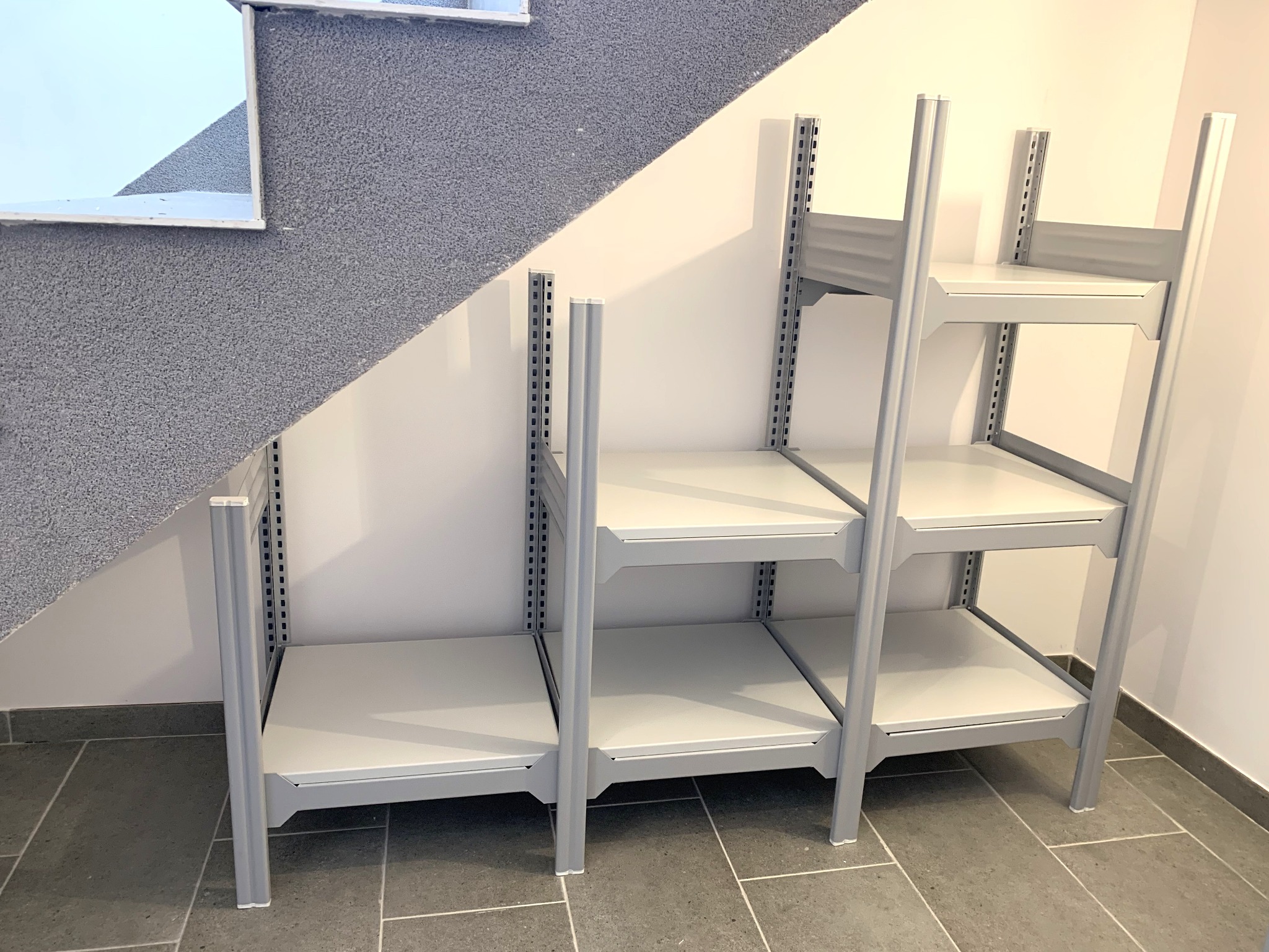 System regałów z blachy ocynkowanej malowanej proszkowo, zamontowany w przestrzeni pod schodami z szarym dywanem. Widoczne perforowane profile pionowe i poziome oraz białe półki.