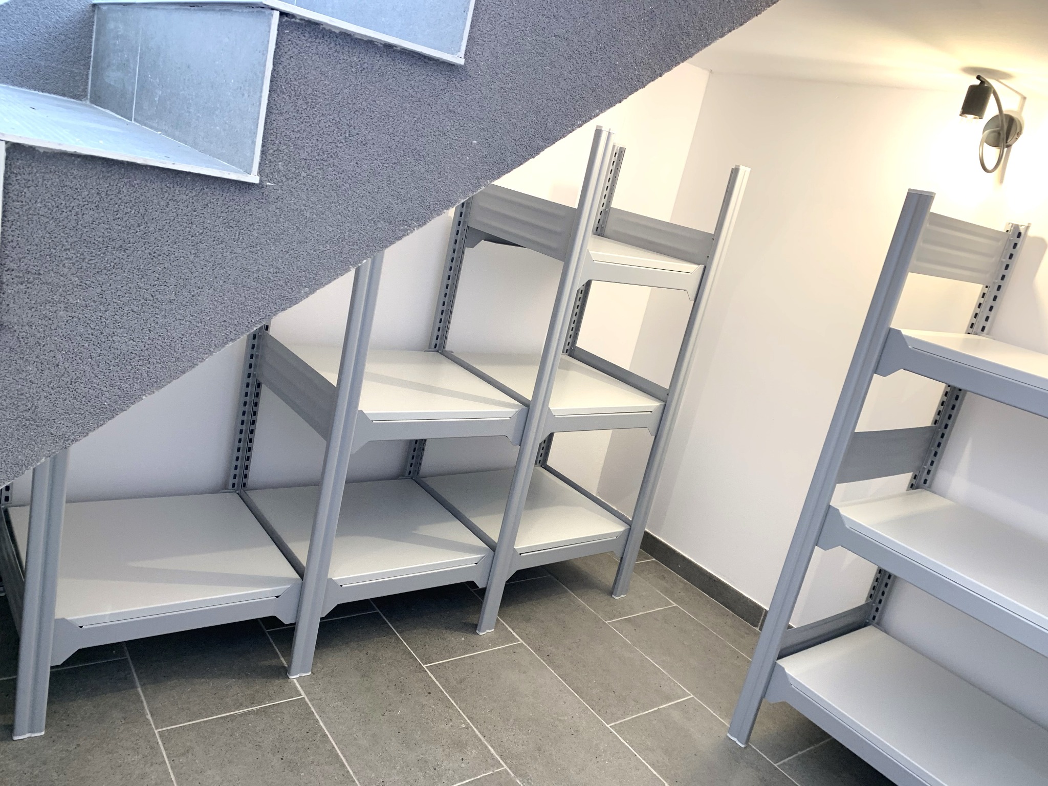 Metalowe regały systemowe z blachy ocynkowanej, malowane proszkowo, zamontowane w przestrzeni pod schodami, widok na szare płytki podłogowe i fragment schodów.