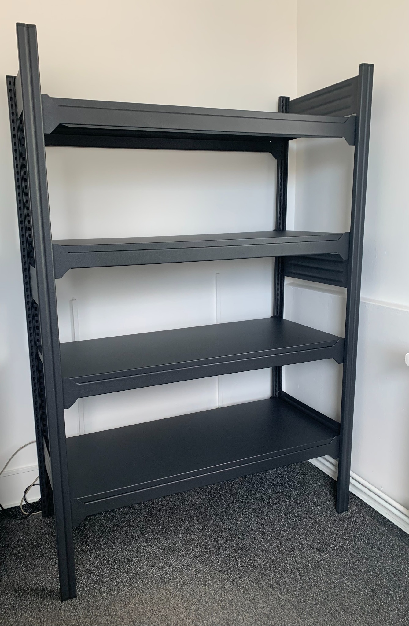Metalowy regał biurowy w kolorze szarym metalik, z czterema półkami z blachy ocynkowanej, ustawiony w rogu pomieszczenia na szarej wykładzinie.