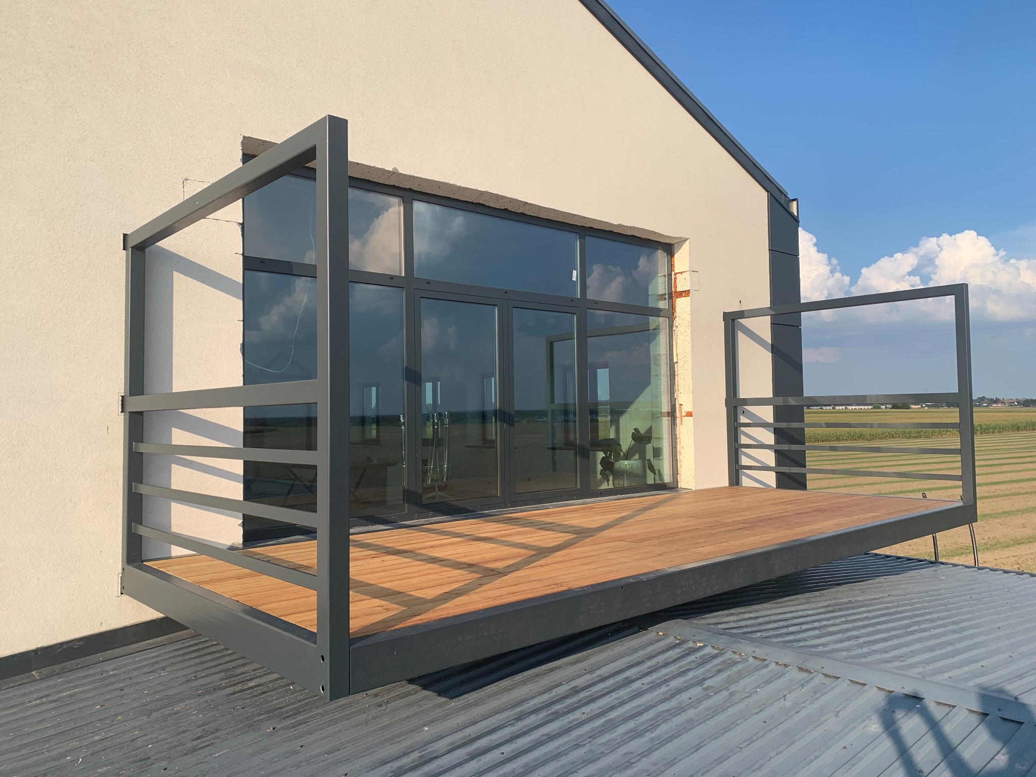 Nowoczesny balkon z drewnianą podłogą i szarą metalową konstrukcją, przylegający do białego budynku z dużymi oknami, widok z dachu.
