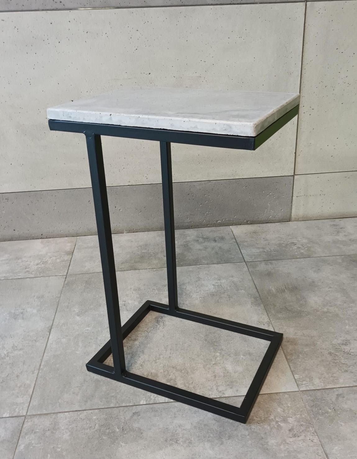 Stolik pomocniczy z blatem z betonu architektonicznego i czarną, metalową podstawą o geometrycznym kształcie na tle szarej, kafelkowanej podłogi i ściany.