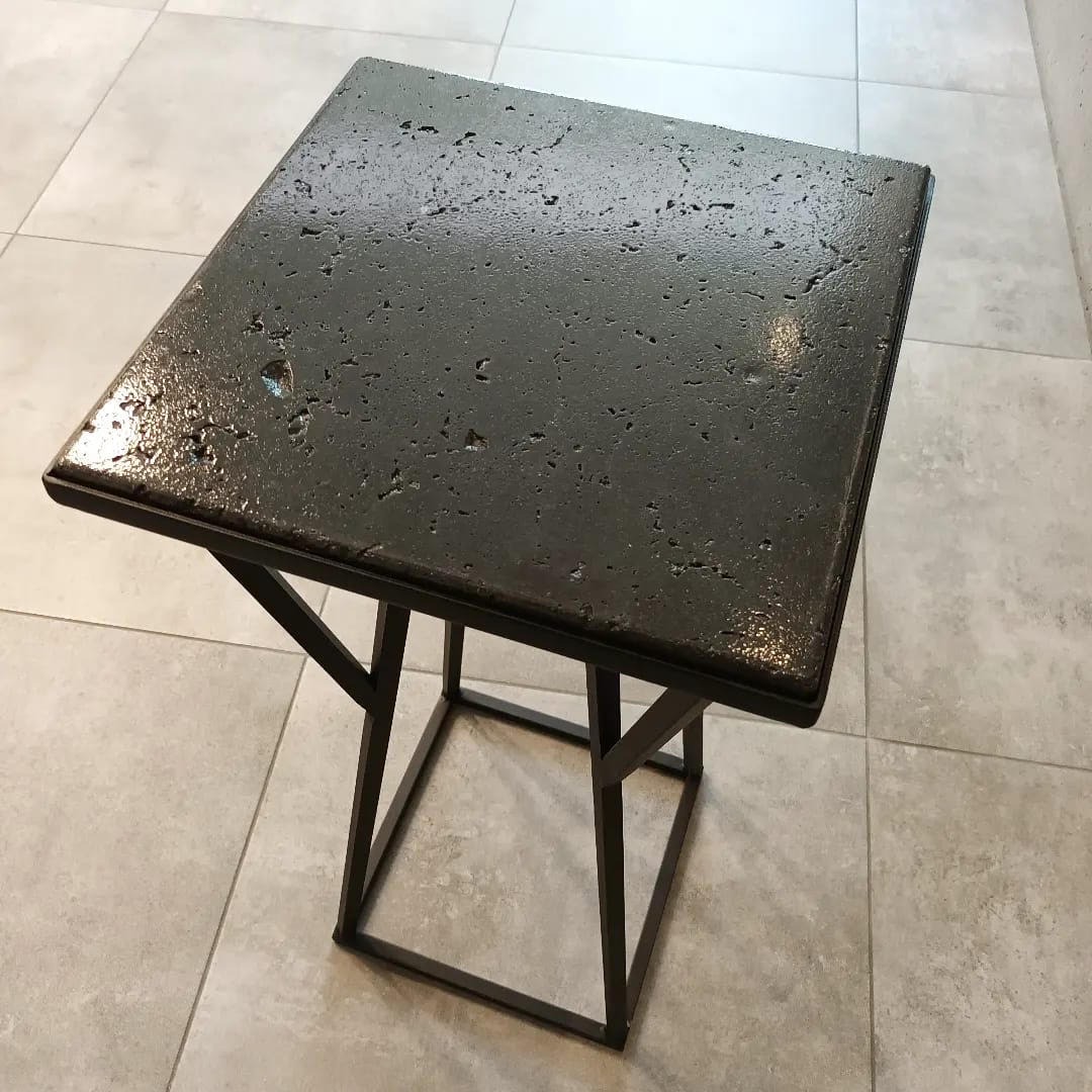 Mały, kwadratowy stolik kawowy z ciemnym, porowatym blatem i metalową podstawą o geometrycznym kształcie, stojący na jasnych, kwadratowych płytkach podłogowych.