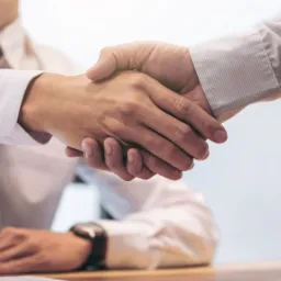 Uścisk dłoni dwóch mężczyzn w koszulach, symbolizujący zawarcie umowy lub partnerstwo biznesowe, z widocznym zegarkiem na nadgarstku jednego z nich i delikatnym oświetleniem bocznym.