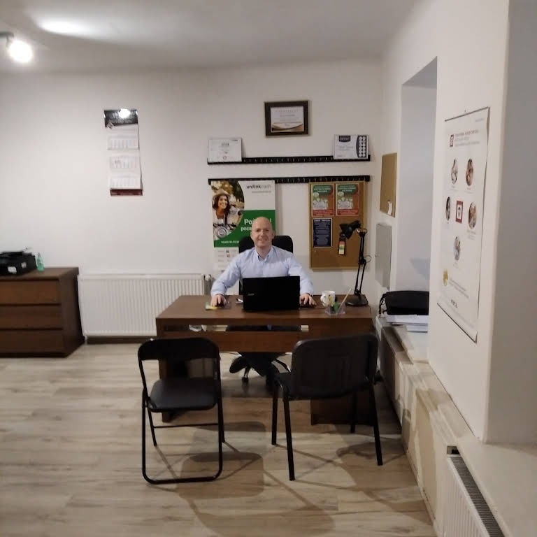 Doradca w jasnym biurze siedzi za biurkiem z laptopem, na ścianie wiszą certyfikaty i plakaty informacyjne.