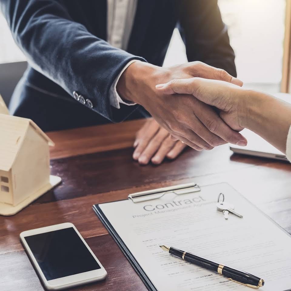 Uścisk dłoni nad umową kredytową, obok miniatura domu, telefon i długopis na drewnianym biurku.