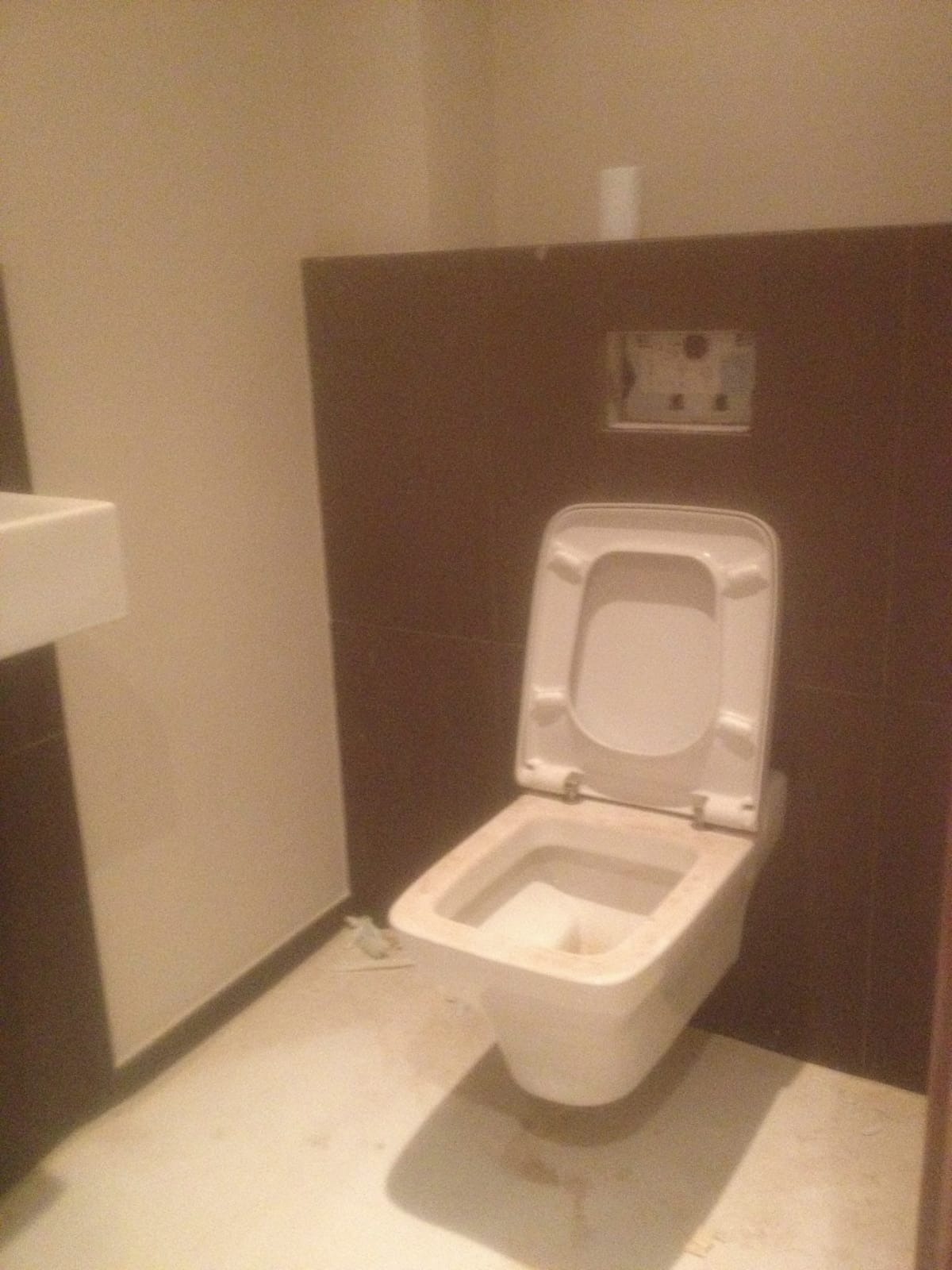 Wisząca toaleta z otwartą klapą, zamontowana na brązowej ścianie w trakcie prac wykończeniowych, widoczne niedociągnięcia i zabrudzenia na podłodze.