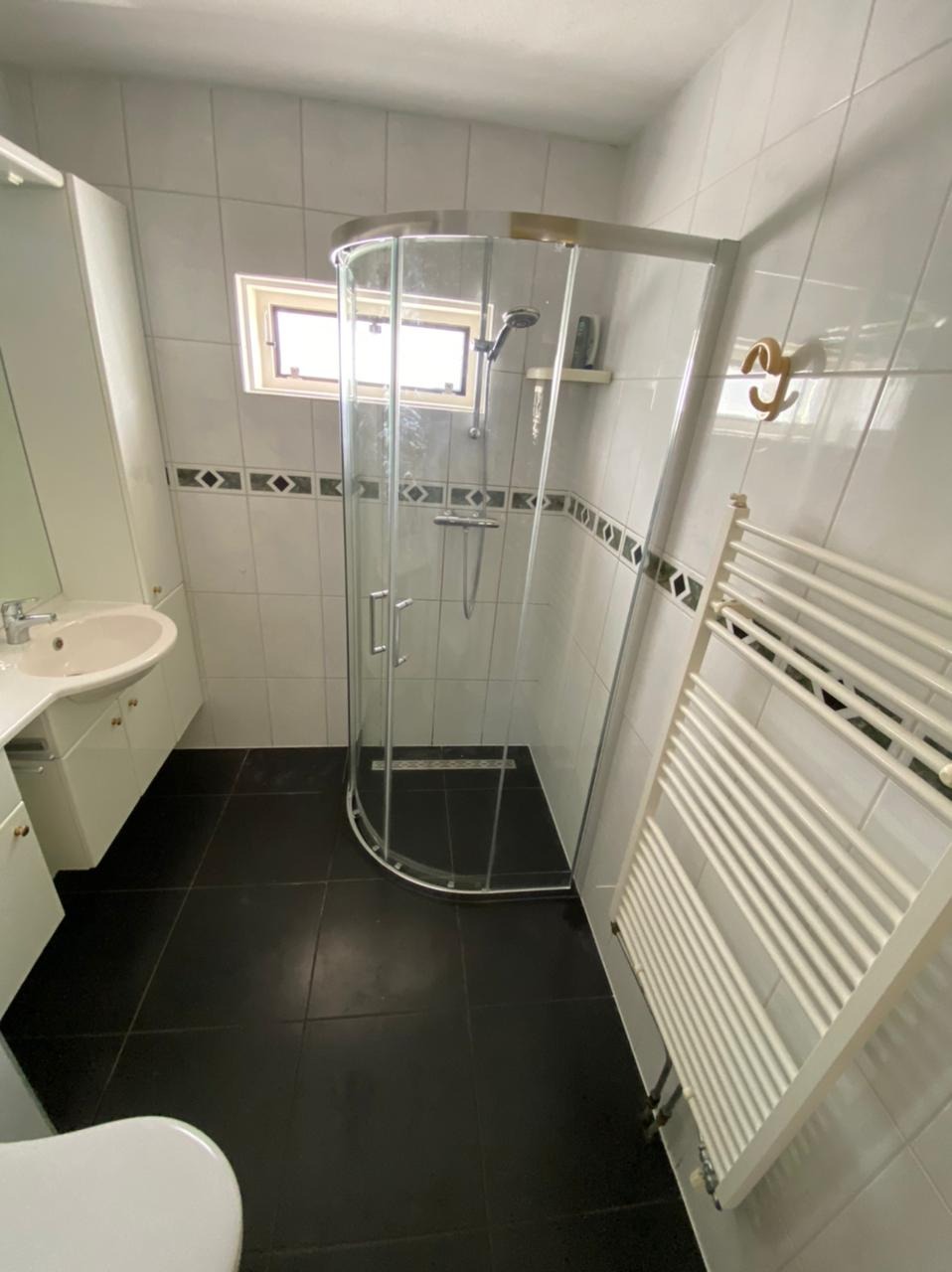 Wykończona łazienka z kabiną prysznicową z zaokrąglonymi szybami, białą umywalką z szafką, grzejnikiem drabinkowym i czarną podłogą z płytek.