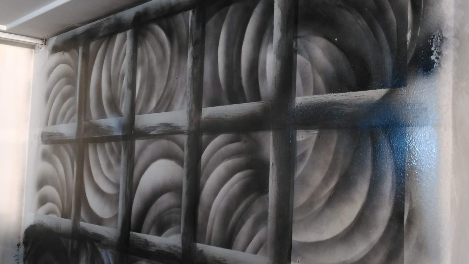 Abstrakcyjna dekoracja ściany w odcieniach szarości z motywem przypominającym spiralne wzory i kratownicę, wykonana techniką malarską z lekkimi niedociągnięciami i drobnymi plamkami farby.