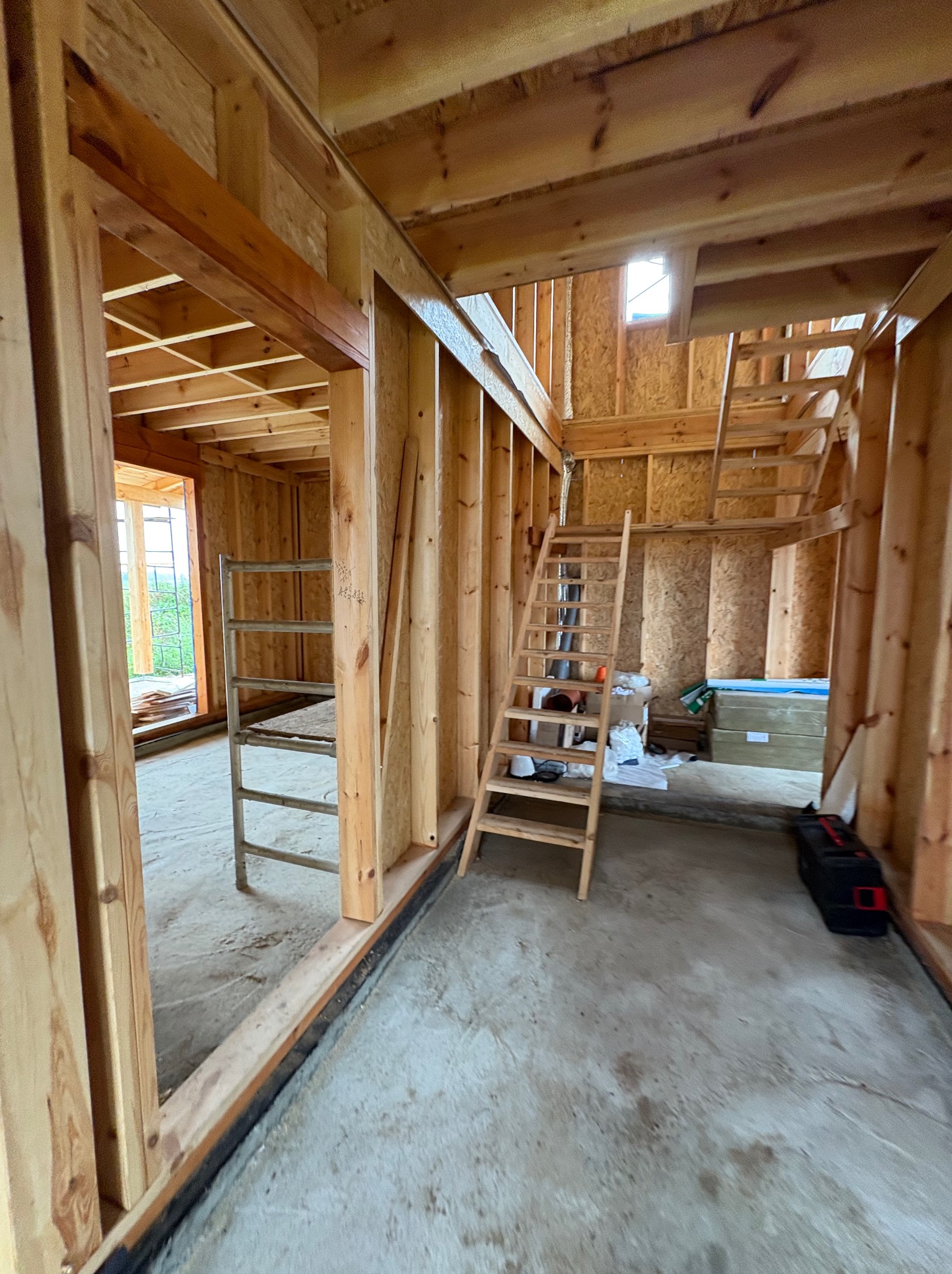 Wnętrze domu w trakcie budowy: drewniana konstrukcja szkieletowa, schody na piętro, widoczne narzędzia i materiały budowlane. Surowy stan, przygotowanie do dalszych prac.