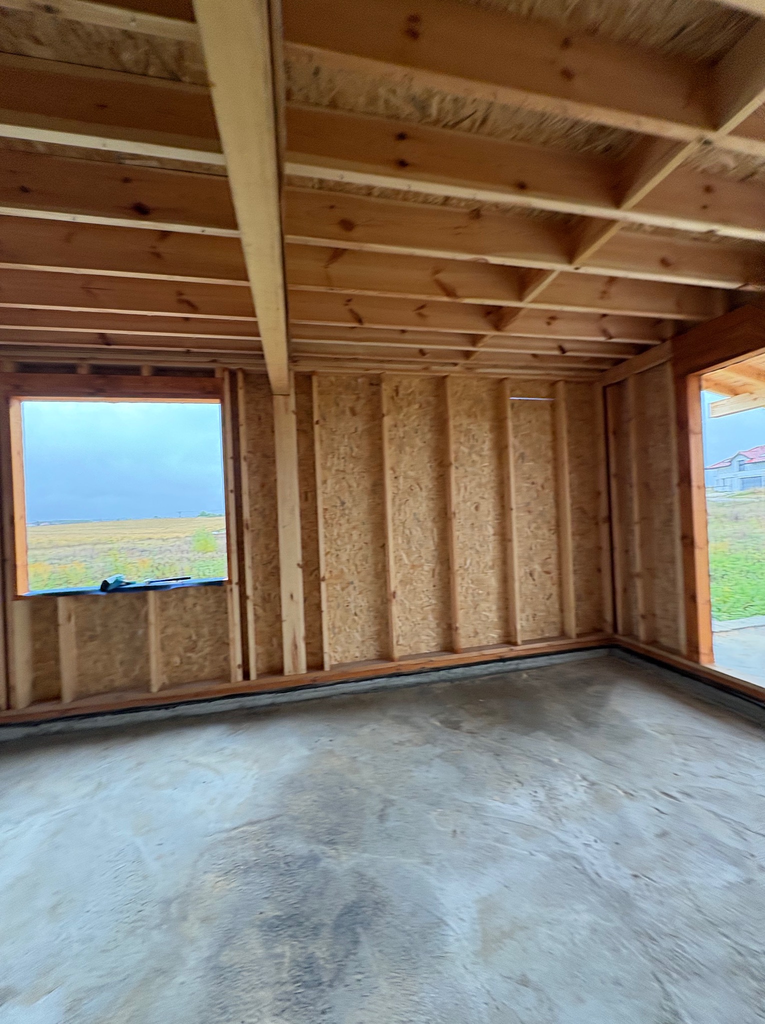 Surowy stan wnętrza domu w trakcie budowy: drewniana konstrukcja ścian, stropu i otwór okienny z widokiem na pole. Betonowa posadzka w szarym odcieniu.