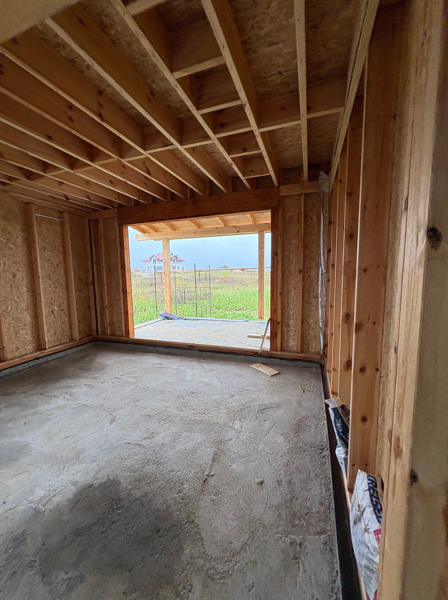 Surowy stan wnętrza budynku w trakcie budowy: drewniana konstrukcja szkieletowa ścian i stropu, wylewka betonowa podłogi, widok na zewnątrz przez otwór okienny.
