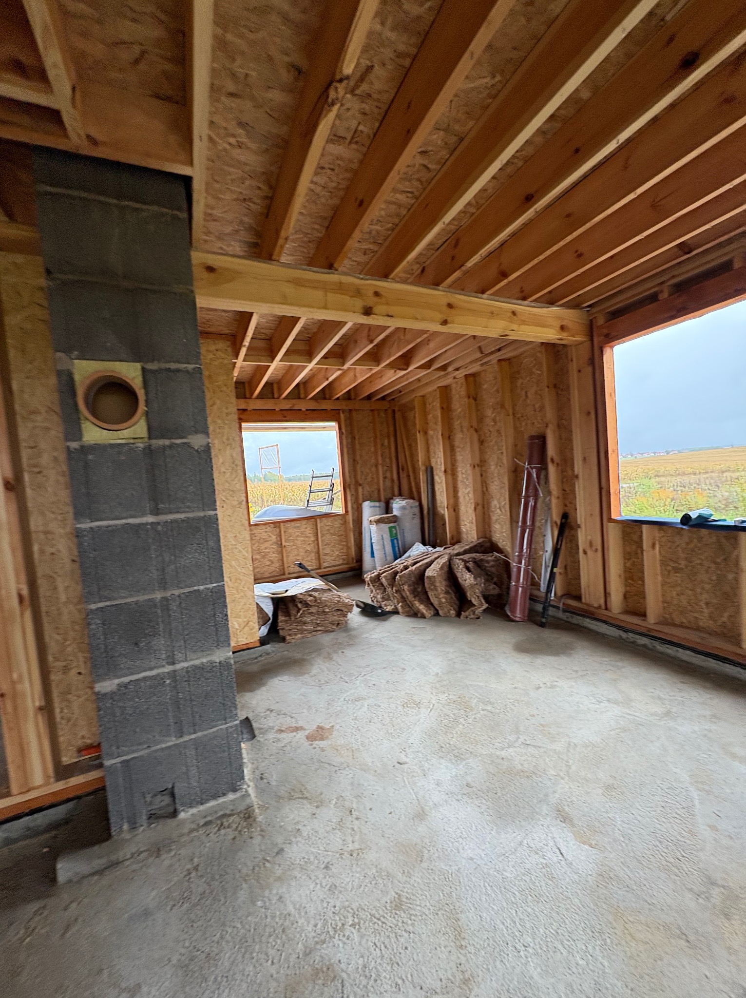 Wnętrze w trakcie budowy: surowy beton, drewniana konstrukcja szkieletowa, izolacja, komin z pustaków. Widok na pole przez okno. Etap prac wykończeniowych.