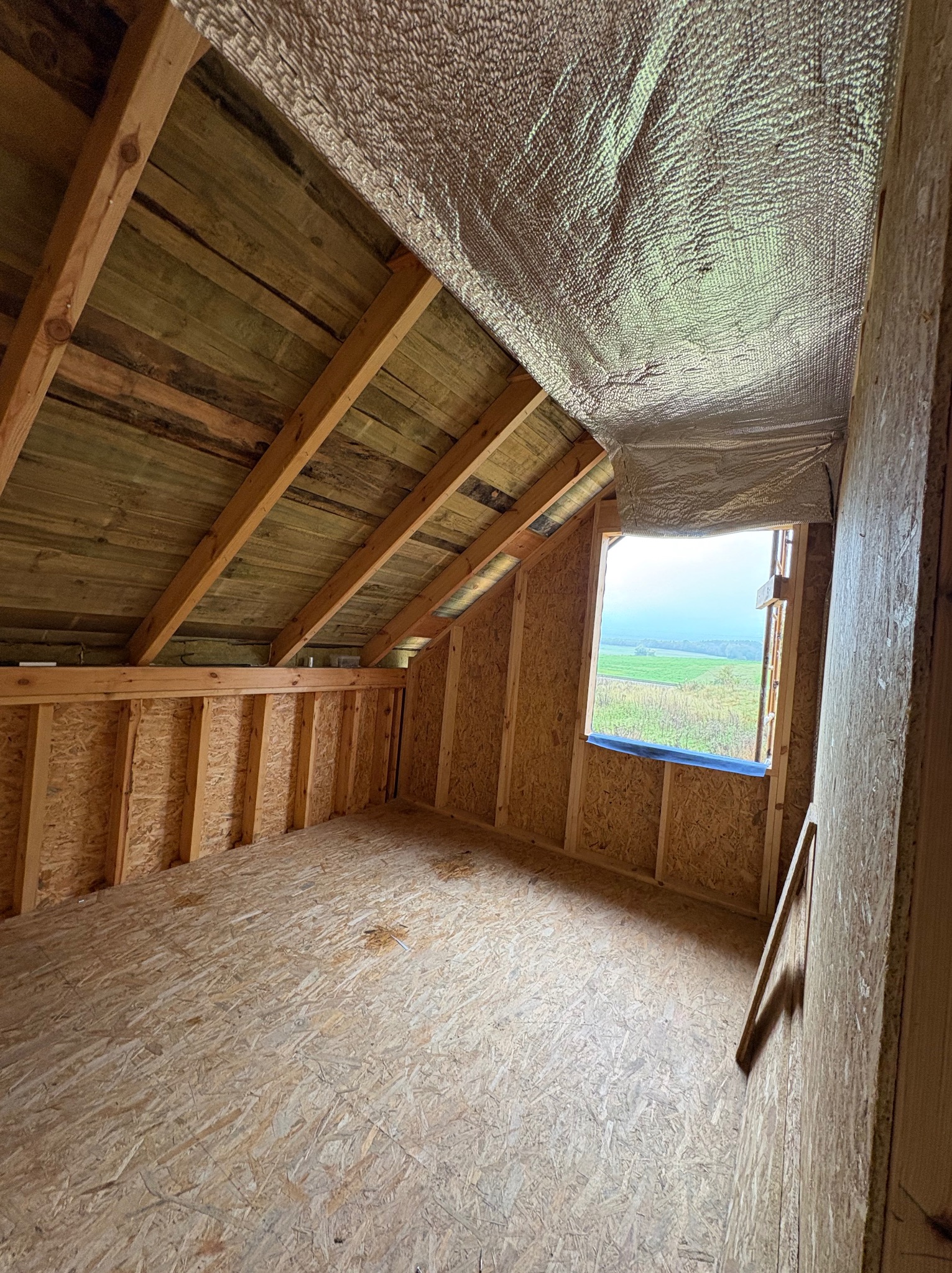 Surowe wnętrze poddasza z drewnianą konstrukcją, płytami OSB na ścianach i podłodze, oraz folią izolacyjną pod dachem. Widoczne okno z widokiem na zewnątrz.