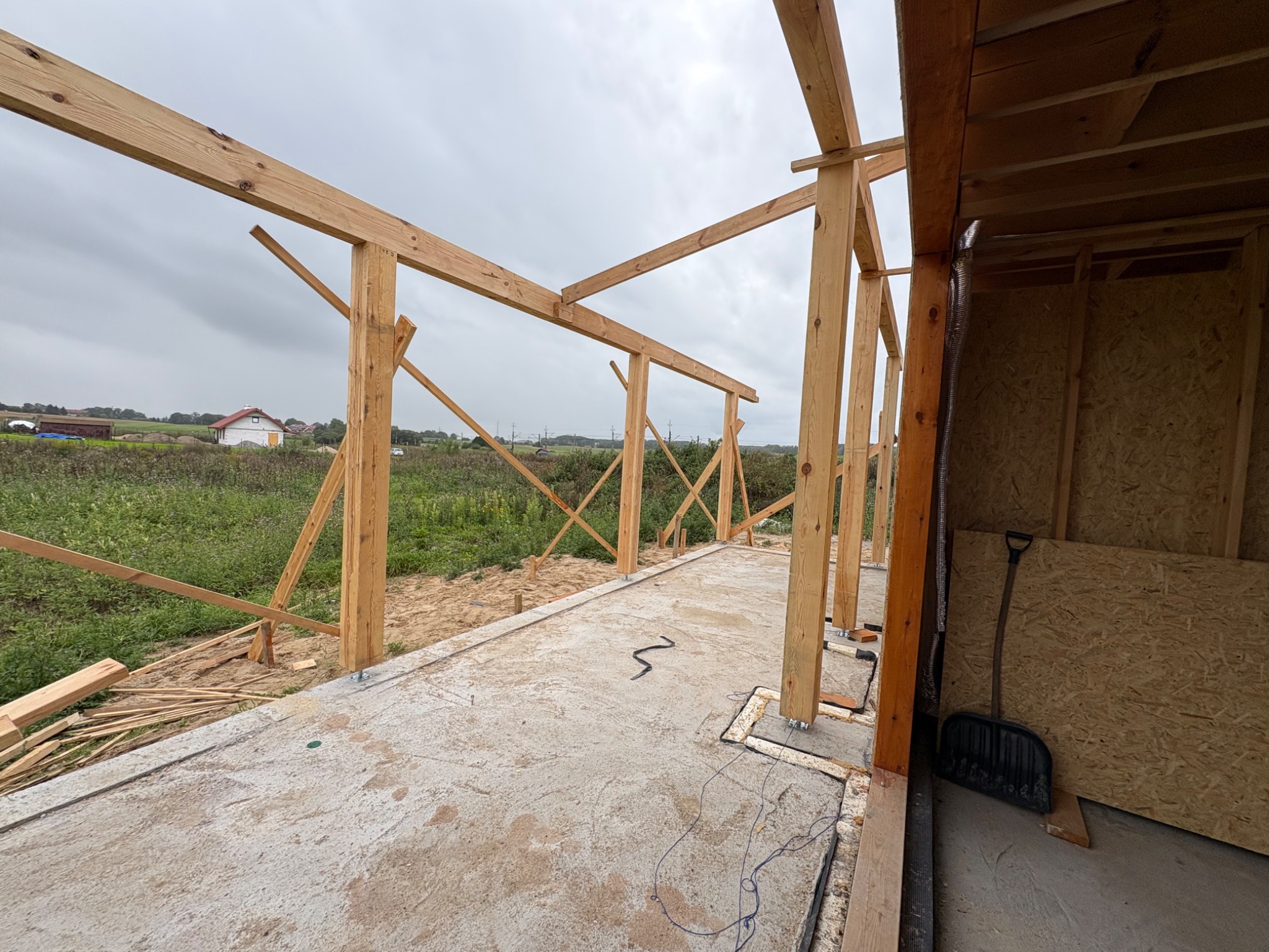 Surowa konstrukcja drewniana budynku w trakcie budowy, widok z wnętrza. Szare niebo w tle. Na podłodze leży łopata.
