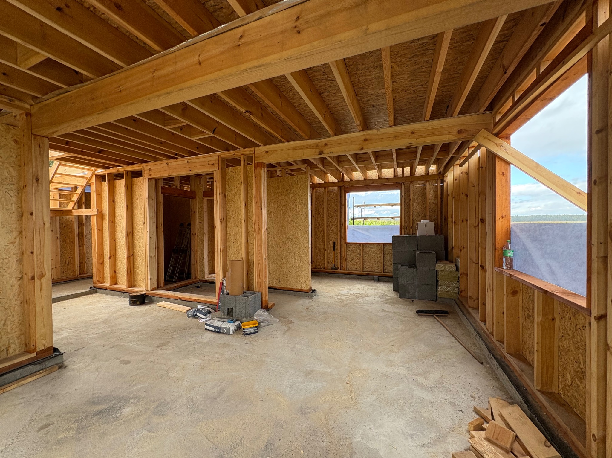 Surowy stan domu w trakcie budowy: drewniana konstrukcja szkieletowa, betonowa podłoga, materiały budowlane wewnątrz. Widok na okno i elementy konstrukcyjne.