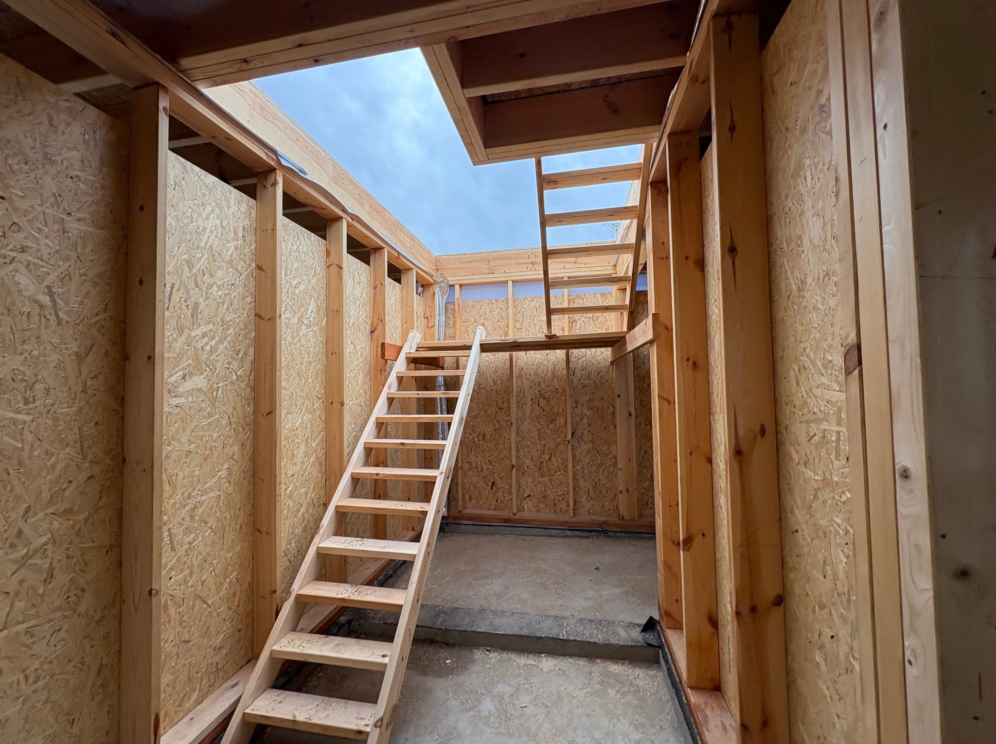 Wnętrze w trakcie budowy: drewniana konstrukcja ścian z płyt OSB, schody prowadzące do otworu w suficie, widoczne niebo. Surowy stan, etap prac wykończeniowych.
