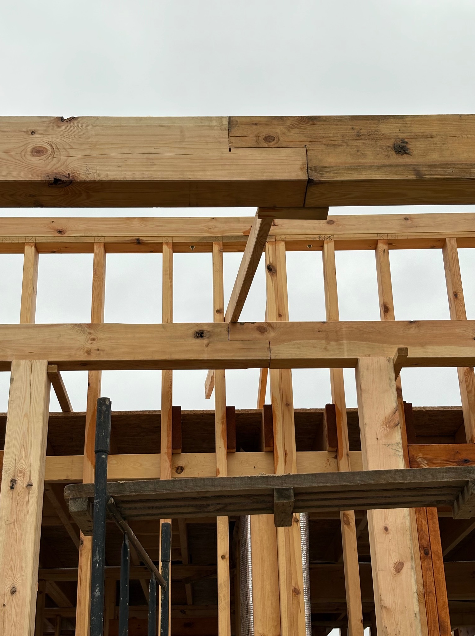 Surowa drewniana konstrukcja szkieletowa domu w trakcie budowy, widok z dołu na belki, słupy i stężenia pod jasnym niebem. Widoczne elementy rusztowania.