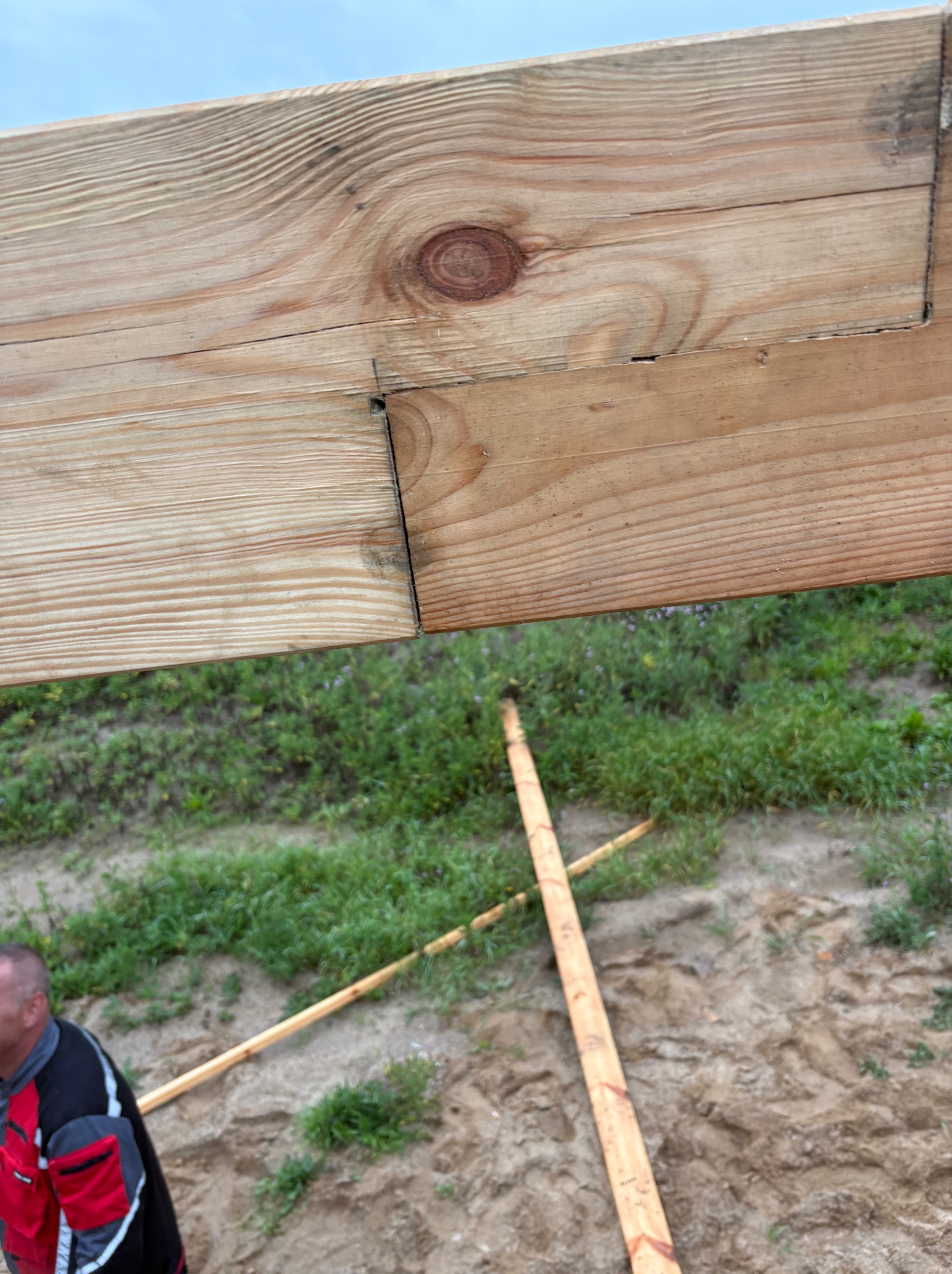 Fragment drewnianej konstrukcji więźby dachowej z widocznym połączeniem elementów, na tle trawy i piasku. Widoczny fragment osoby w czerwonej kurtce.
