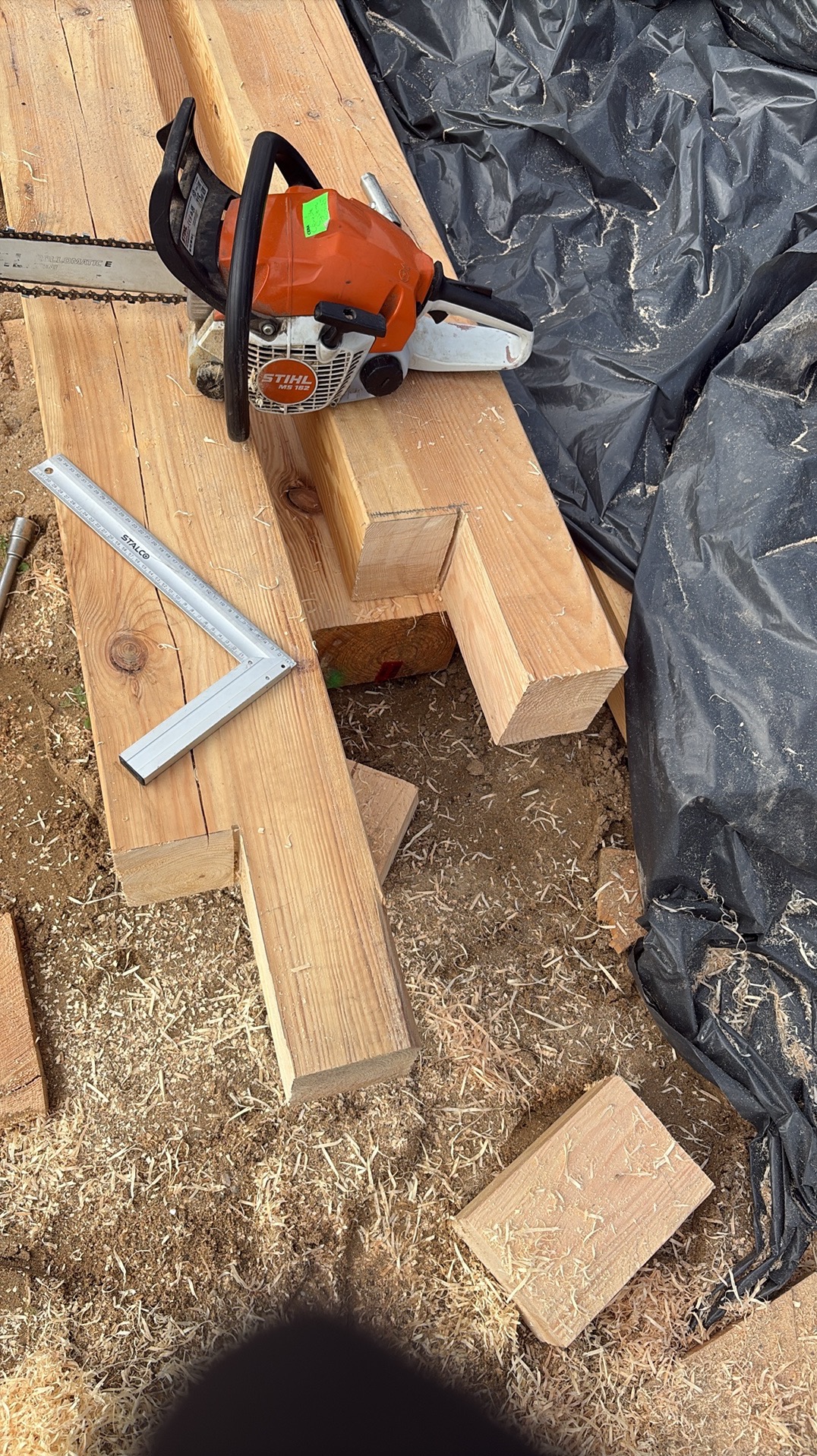 Piła spalinowa Stihl na drewnianych belkach z wyciętymi fragmentami, obok kątownik stolarski. Wióry i czarna folia w tle. Przygotowanie konstrukcji drewnianej.