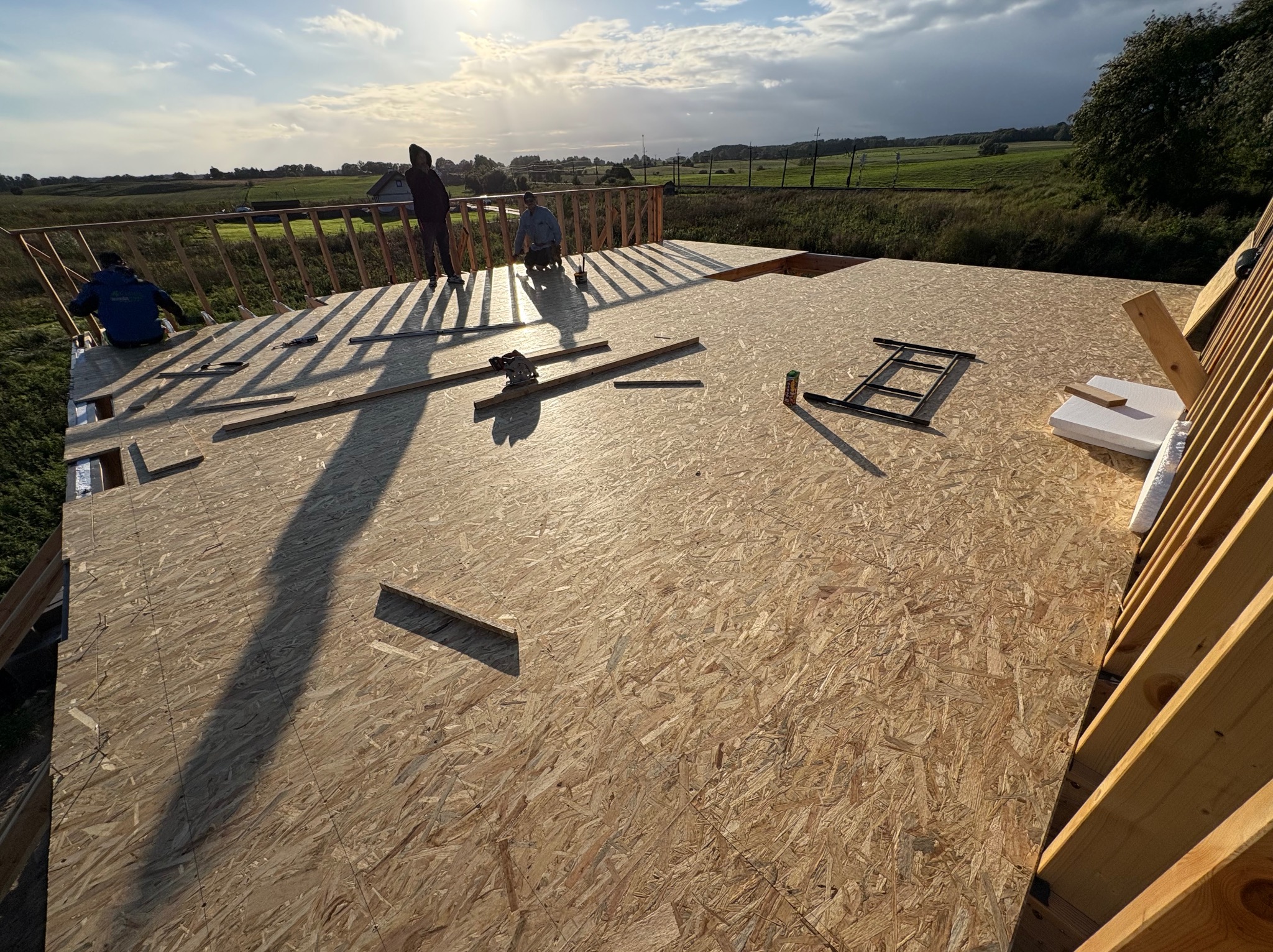 Montaż płyt OSB na konstrukcji dachu domu jednorodzinnego. Widoczne narzędzia, elementy konstrukcyjne i pracownicy w tle. Słoneczny dzień, widok na okolicę.