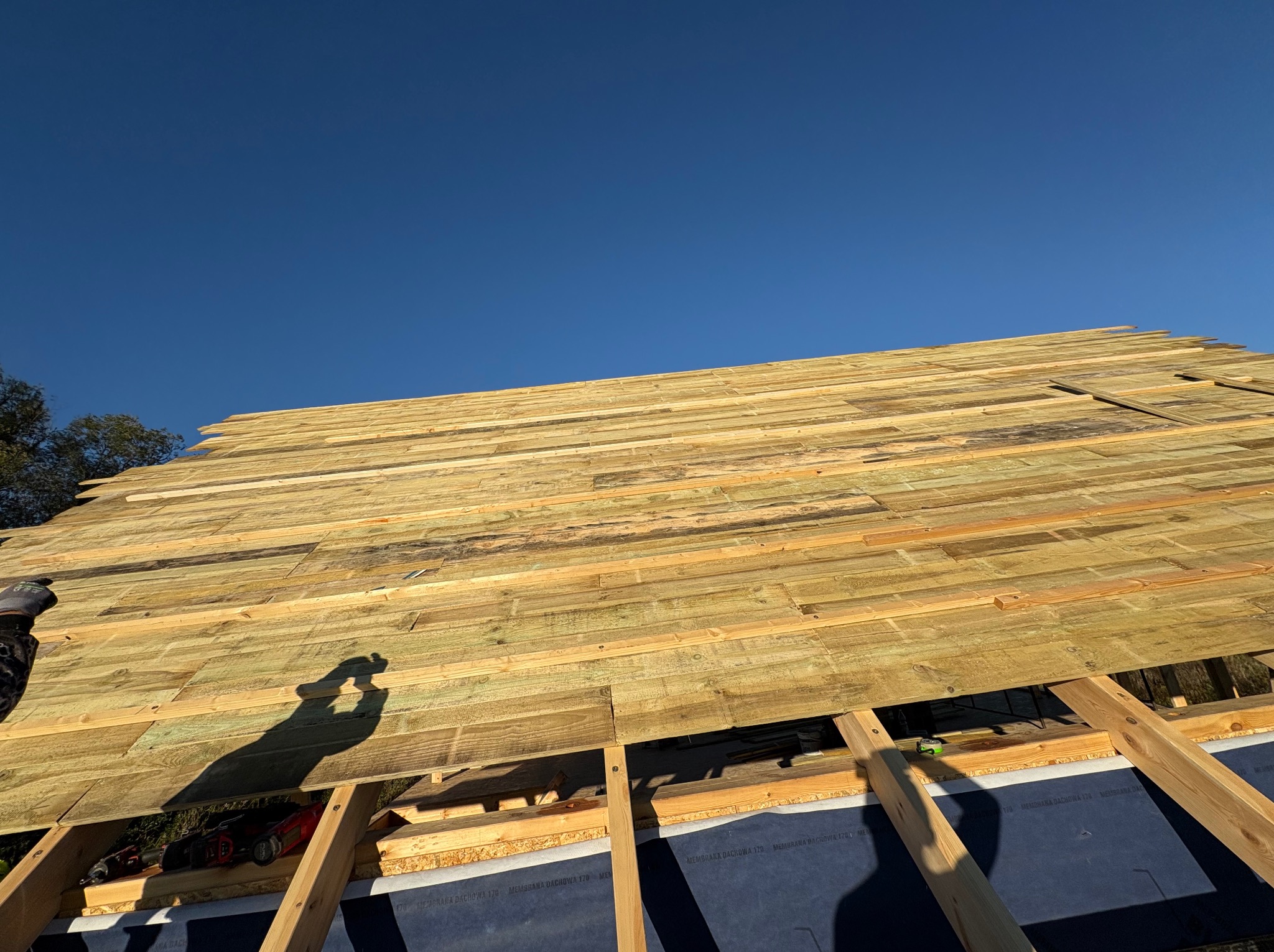 Deskowanie dachu z widoczną membraną dachową i narzędziami. Ujęcie z dołu, na pierwszym planie cień osoby. Błękitne niebo w tle.