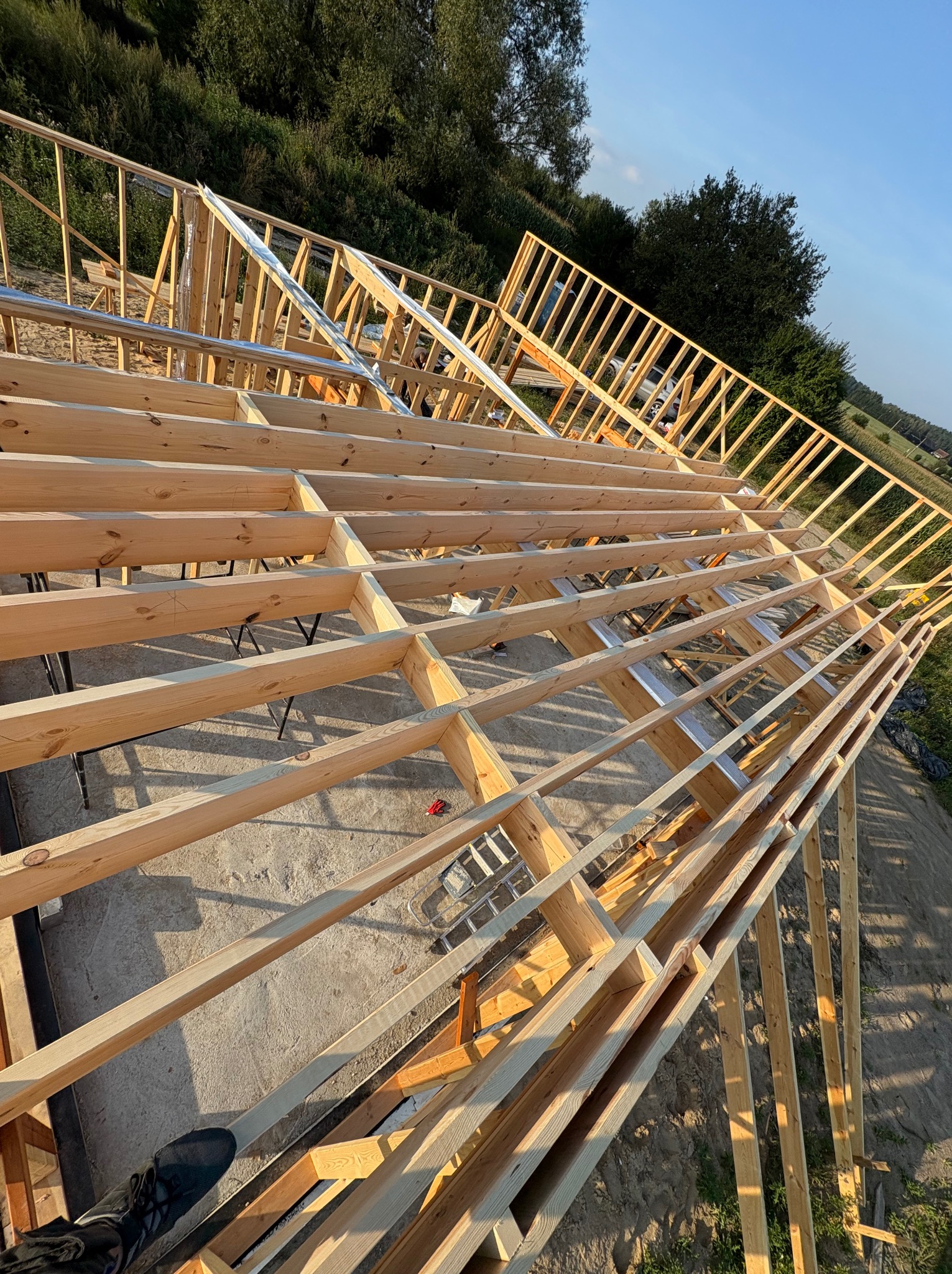 Konstrukcja drewniana dachu w trakcie budowy, widok z góry. Elementy więźby dachowej z jasnego drewna na tle zieleni i błękitnego nieba. Widoczne detale łączeń i mocowań.