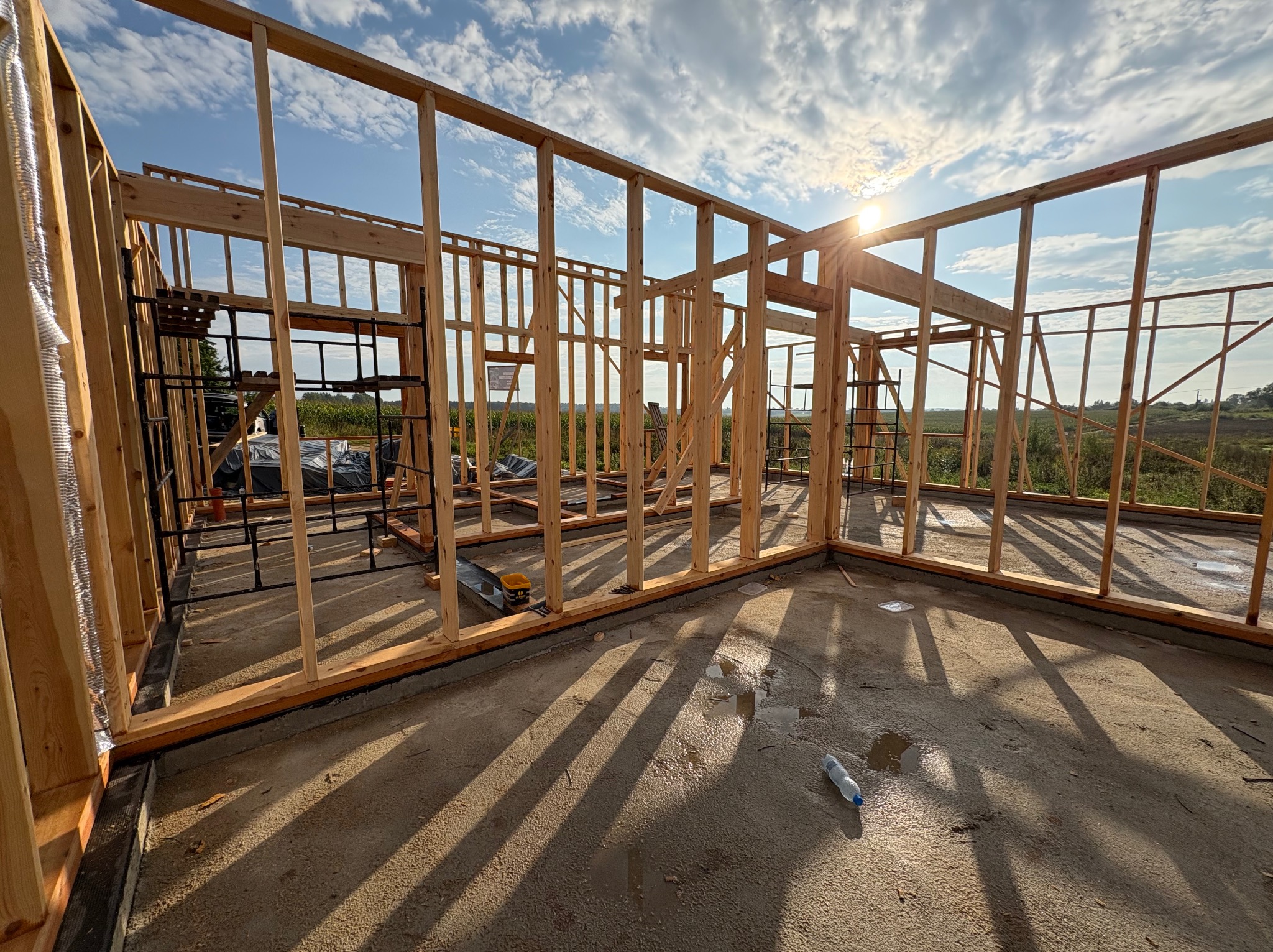 Surowa konstrukcja szkieletowa domu w słoneczny dzień. Widoczne drewniane belki, podłoga betonowa z kałużami i butelka wody. W tle zieleń i błękitne niebo.