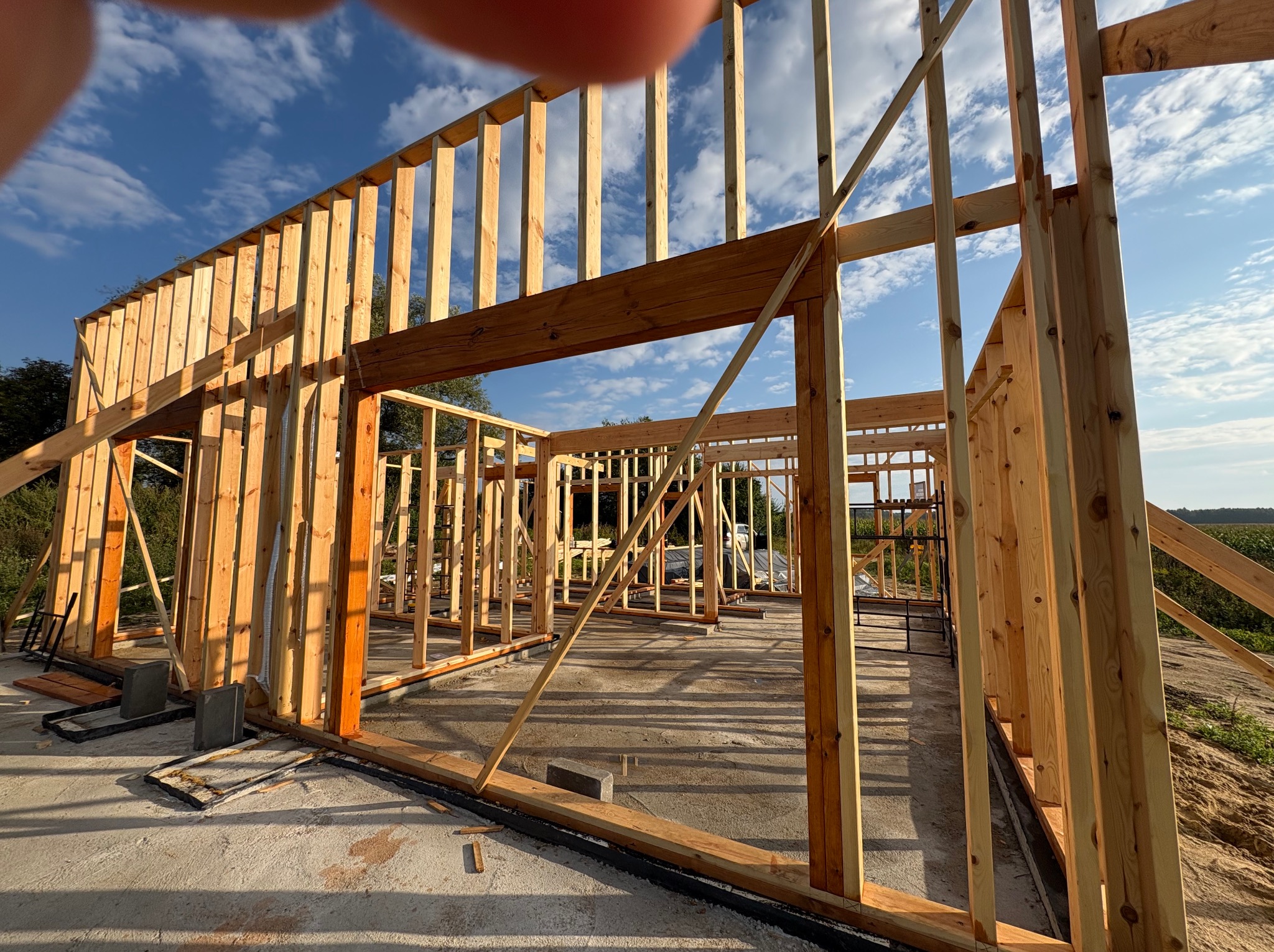 Surowa konstrukcja szkieletowa budynku w trakcie budowy, widoczne drewniane belki i słupy na betonowej podstawie, błękitne niebo w tle. Częściowo zasłonięte ujęcie palcem.