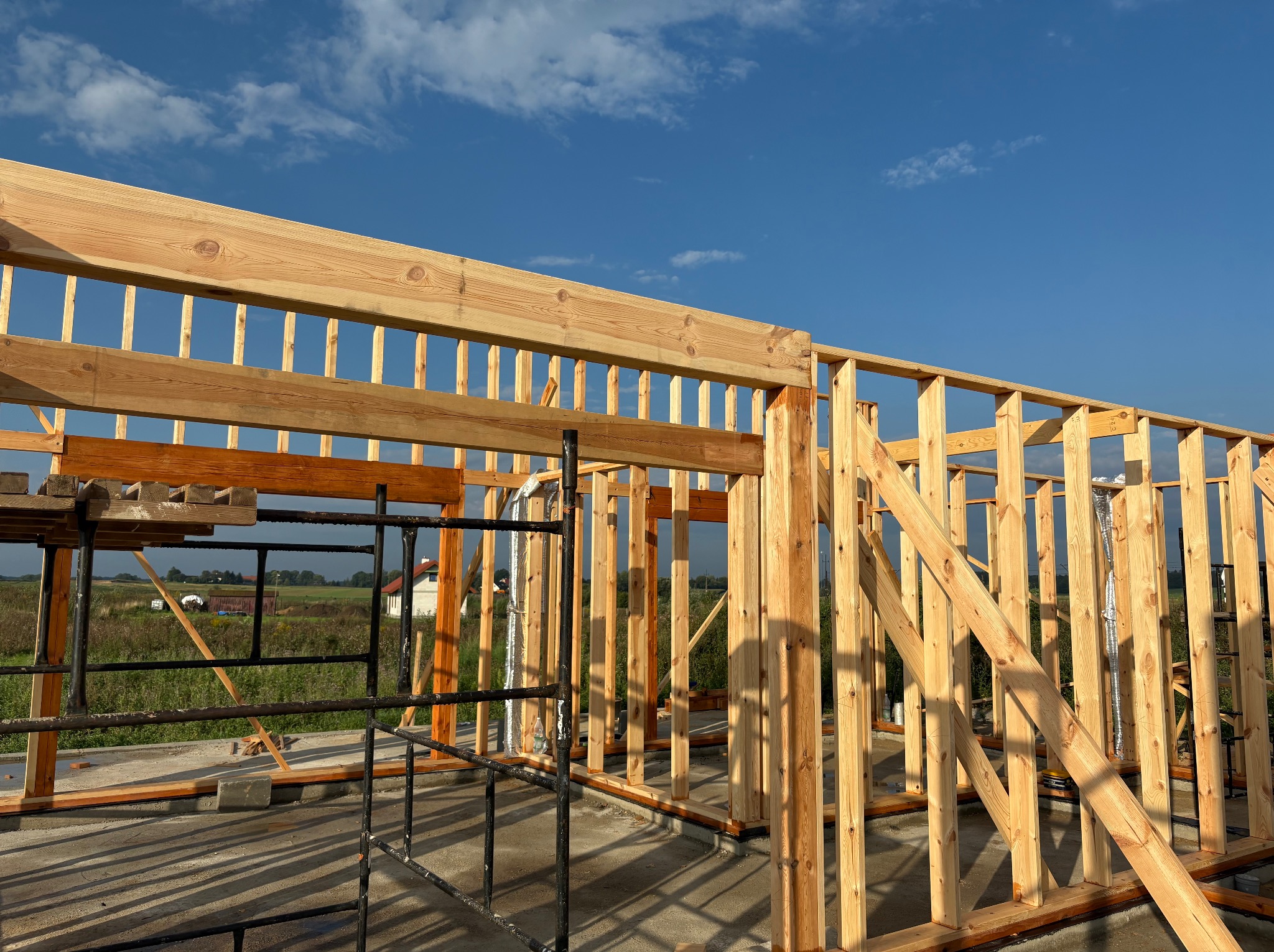 Surowa drewniana konstrukcja szkieletowa domu w trakcie budowy, widok z zewnątrz na tle błękitnego nieba i zieleni. Widoczne rusztowania i elementy zabezpieczeń.