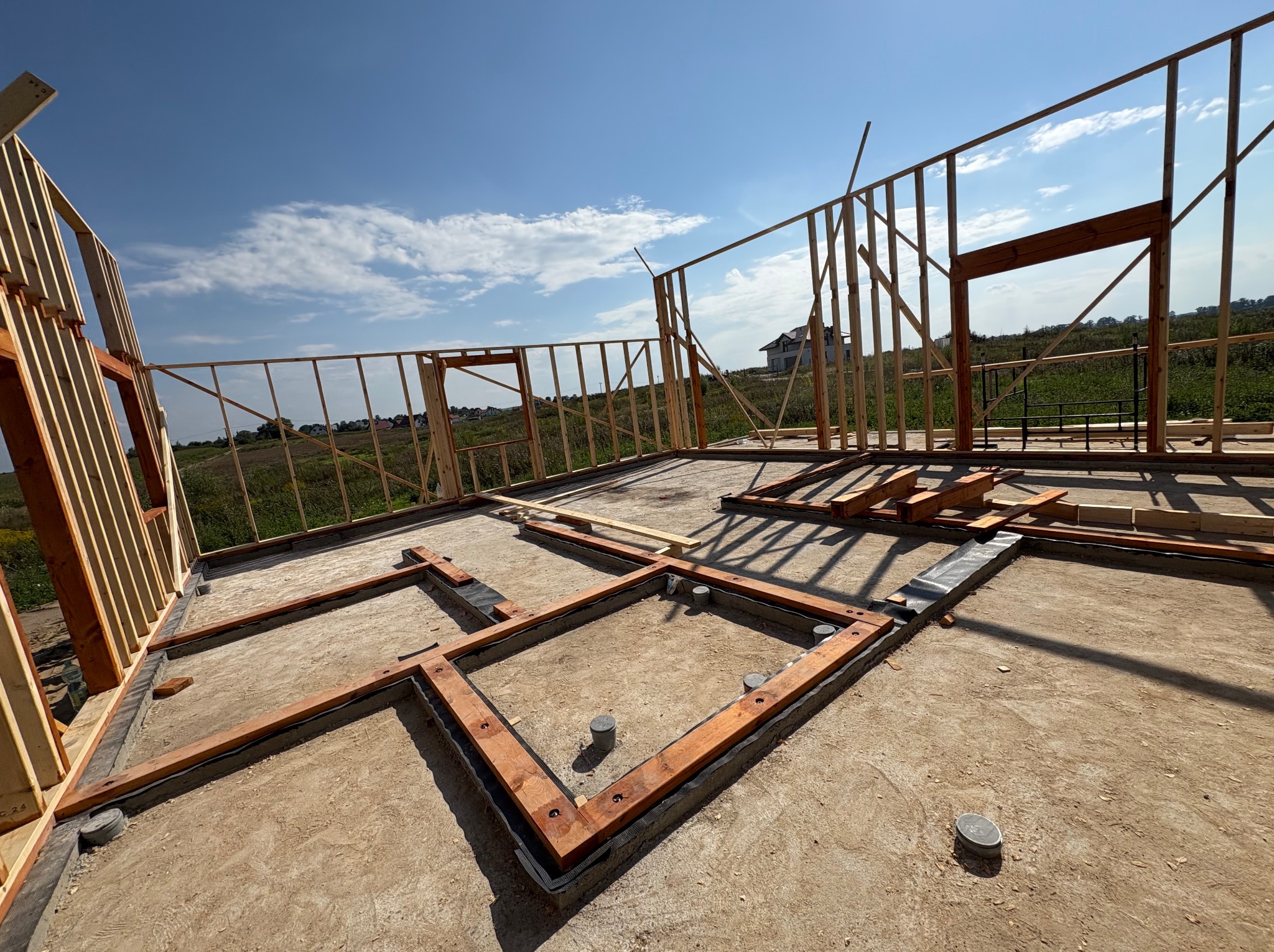 Surowy stan budowy domu: drewniany szkielet ścian i fundamentów na betonowej płycie, widok na otwarty krajobraz i błękitne niebo z chmurami.
