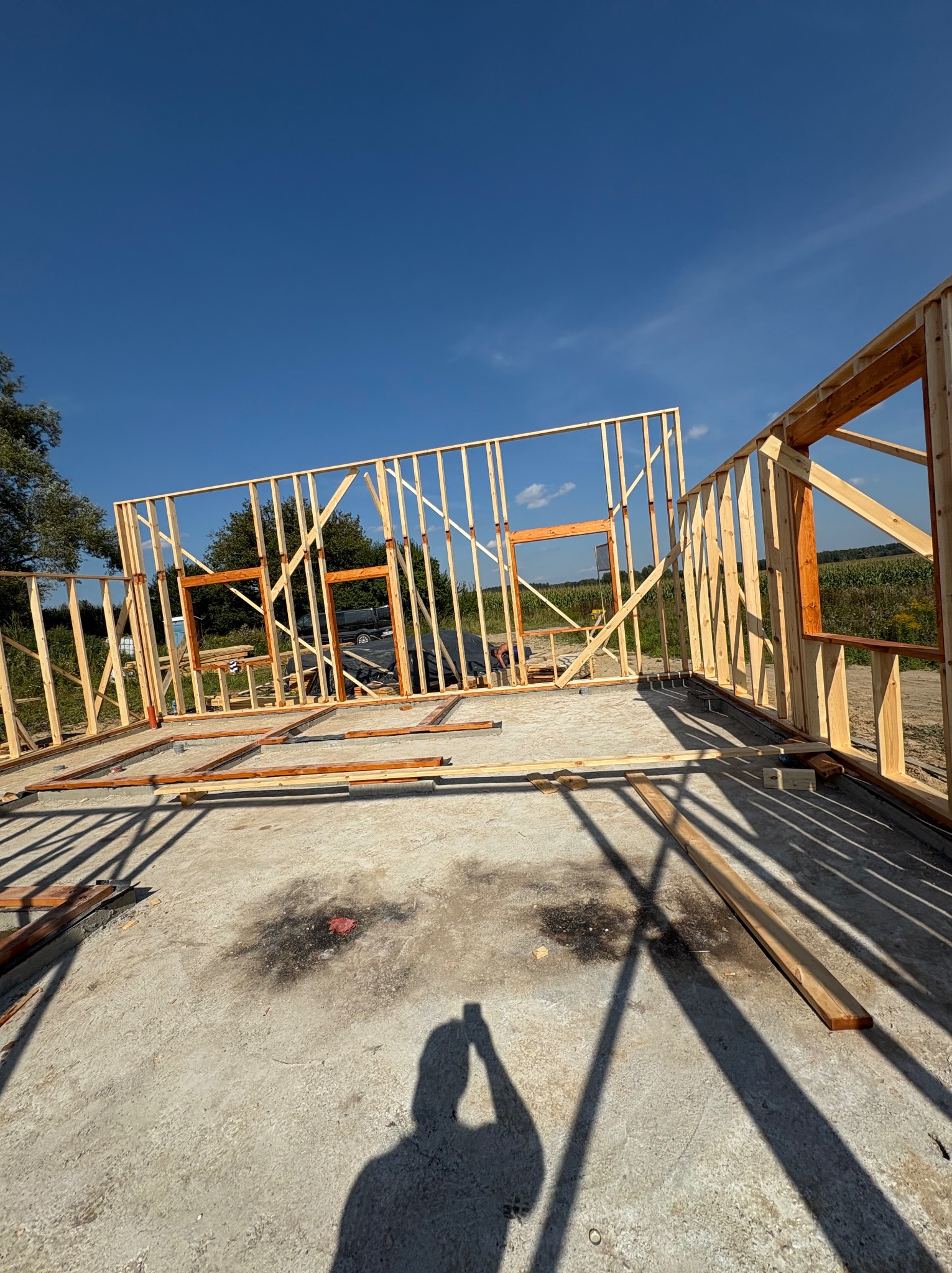 Wznoszenie drewnianej konstrukcji szkieletowej budynku z widocznymi otworami okiennymi na betonowej płycie fundamentowej, w tle błękitne niebo i zieleń drzew.