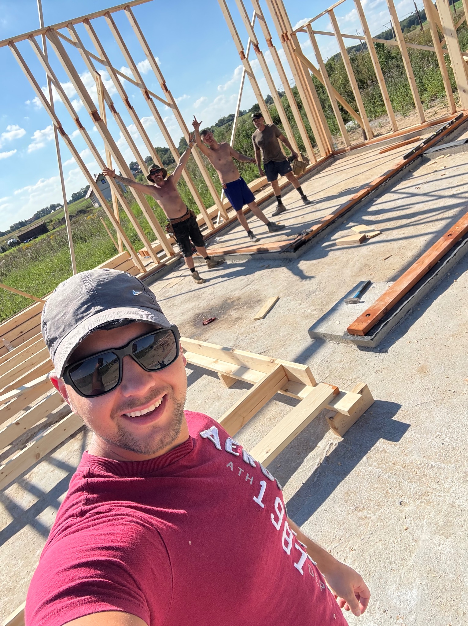 Ekipa budowlana świętuje postęp prac przy wznoszeniu drewnianej konstrukcji szkieletowej domu. Widoczny fundament i częściowo wzniesione ściany. Radosna atmosfera.