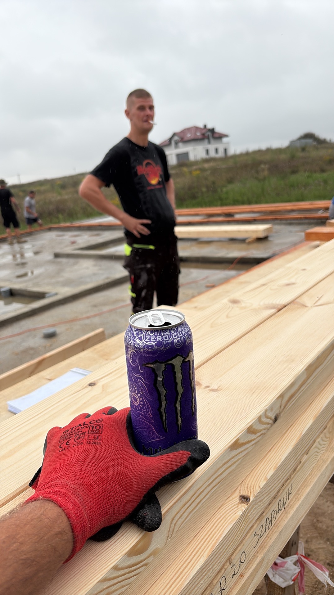 Konstrukcja drewniana w trakcie budowy domu, puszka napoju energetycznego i rękawica robocza na pierwszym planie, w tle pracownik budowlany i zarys domu.