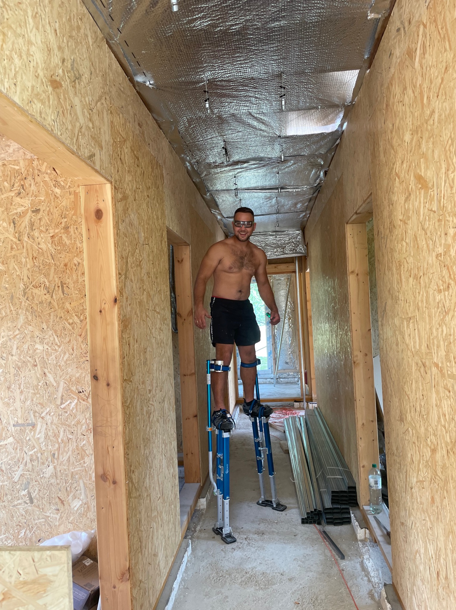 Pracownik na szczudłach montuje płyty OSB wewnątrz budynku. Widoczne ocieplenie sufitu folią aluminiową. W tle elementy konstrukcyjne i narzędzia.