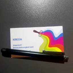 Wizytówka firmy KREON z grafiką pędzla malarskiego rozprowadzającego kolorowe farby, leżąca obok czarnego długopisu na ciemnoszarym tle.