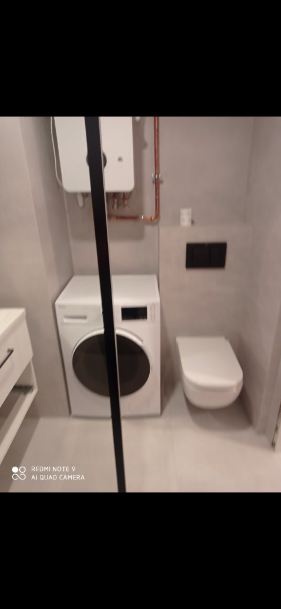 Nowoczesna łazienka z pralką, podwieszaną toaletą i bojlerem, wykończona w szarych płytkach, widok zza kabiny prysznicowej.