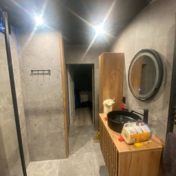 Krys-Bud usługi ogólnobudowlane Krystian Szweda - Nowoczesna łazienka z szarą płytką, drewnianą szafką pod umywalką i okrągłym lustrem z podświetleniem. Widoczny fragment kabiny prysznicowej.