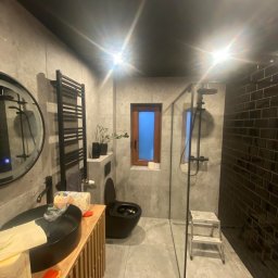 Krys-Bud usługi ogólnobudowlane Krystian Szweda - Nowoczesna łazienka z czarną armaturą: umywalka, grzejnik drabinkowy, toaleta. Ściany wyłożone szarą płytką, kabina prysznicowa ze szklaną ścianą i czarną glazurą.